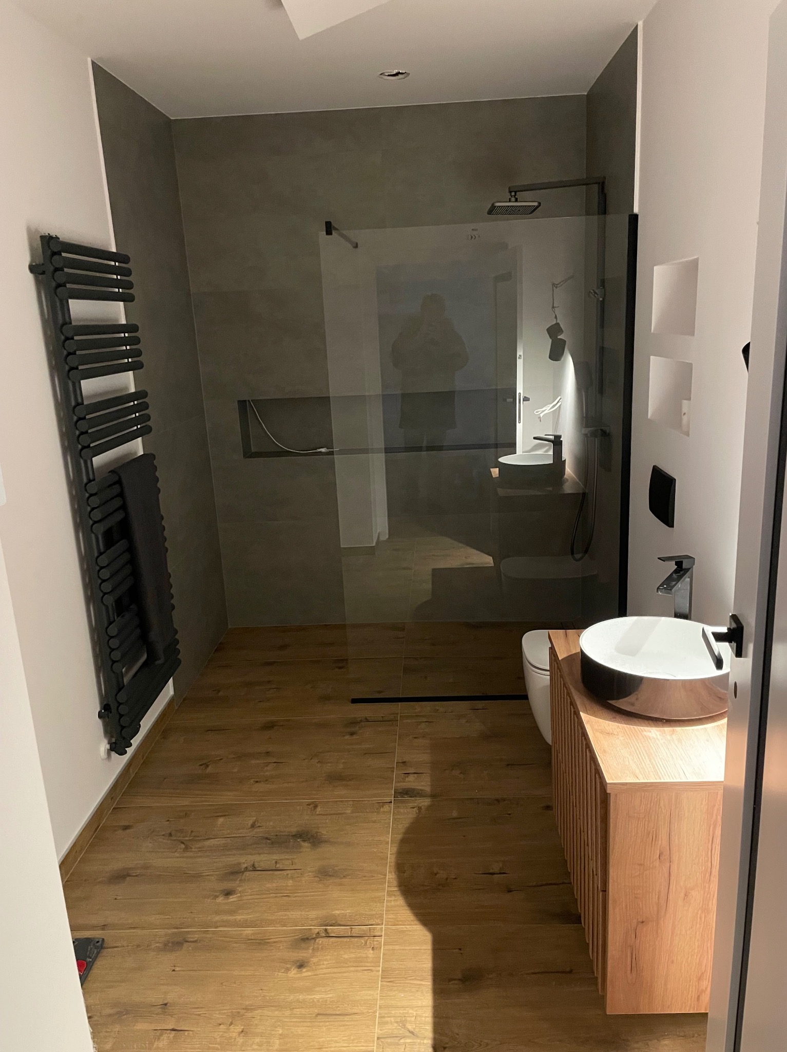 Nowoczesna łazienka z kabiną prysznicową typu walk-in, drewnopodobnymi płytkami podłogowymi i grzejnikiem drabinkowym w kolorze czarnym. Umywalka nablatowa.