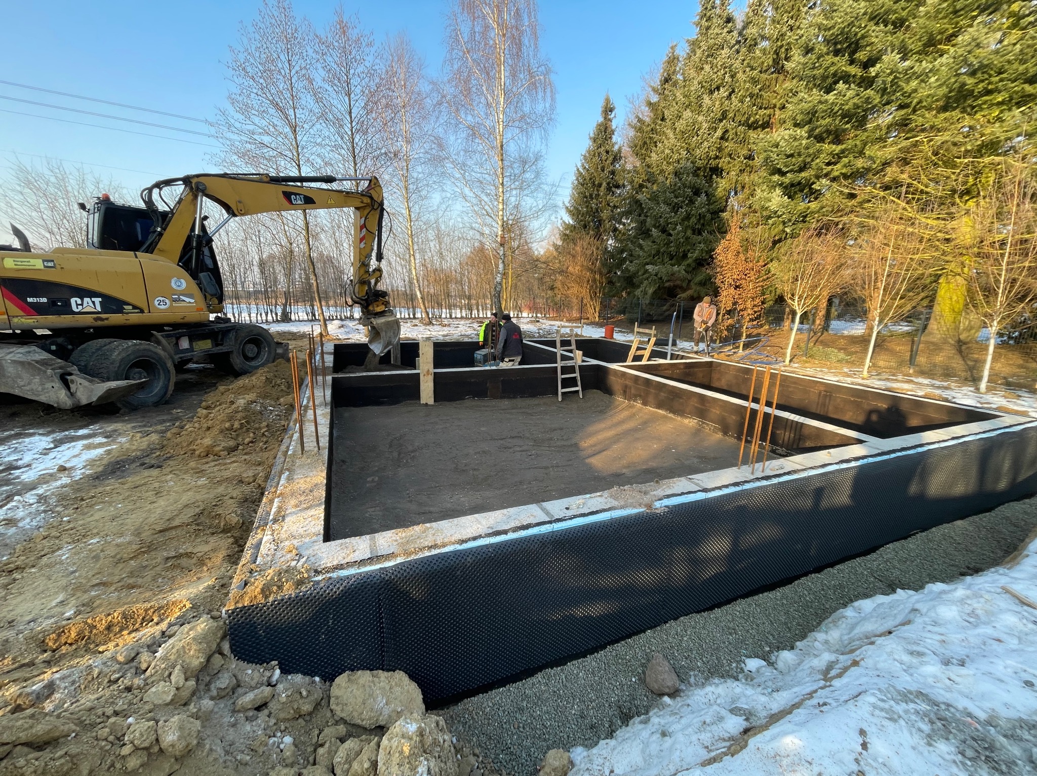 Fundamenty domu w trakcie budowy, z widoczną izolacją przeciwwilgociową i częściowo zasypanym wykopem. Na pierwszym planie ziemia i śnieg, w tle drzewa i błękitne niebo.