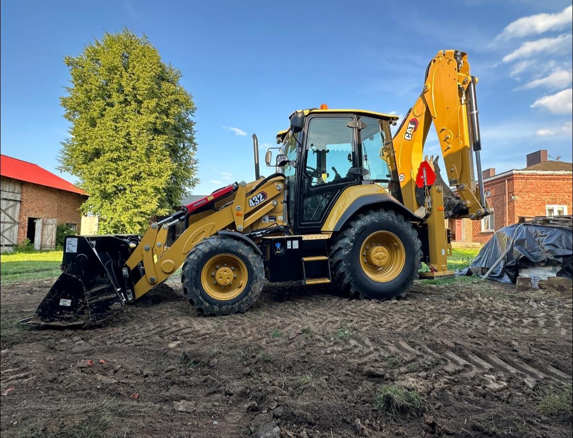 Żółta koparko-ładowarka CAT 432 stoi na ubitej ziemi na tle wiejskiego krajobrazu z drzewem i budynkami z czerwonej cegły.