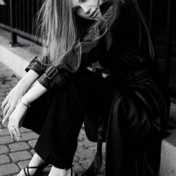 Patrycja Błajdo Fotografia - Czarno-białe zdjęcie kobiety w czarnym płaszczu i spodniach siedzącej na krawężniku przy metalowym ogrodzeniu, z delikatnym uśmiechem i spojrzeniem w obiektyw, włosy opadają na ramiona.