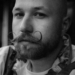 Patrycja Błajdo Fotografia - Czarno-białe portretowe ujęcie mężczyzny z ogoloną głową i charakterystycznymi, zakręconymi wąsami, ubrany w bluzę z zamkiem błyskawicznym.