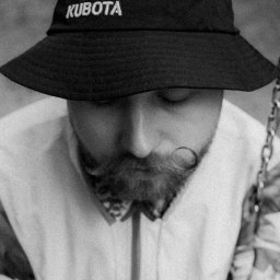 Patrycja Błajdo Fotografia - Portret mężczyzny z brodą i wąsami w czarnym kapeluszu z logo Kubota, czarno-białe zdjęcie z bliska.