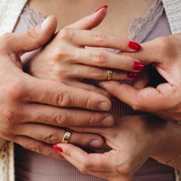 Patrycja Błajdo Fotografia - Zbliżenie dłoni pary z obrączkami ślubnymi; kobiece dłonie z czerwonym manicure trzymane w męskich dłoniach, delikatna bransoletka na nadgarstku kobiety, ażurowy sweter w tle.