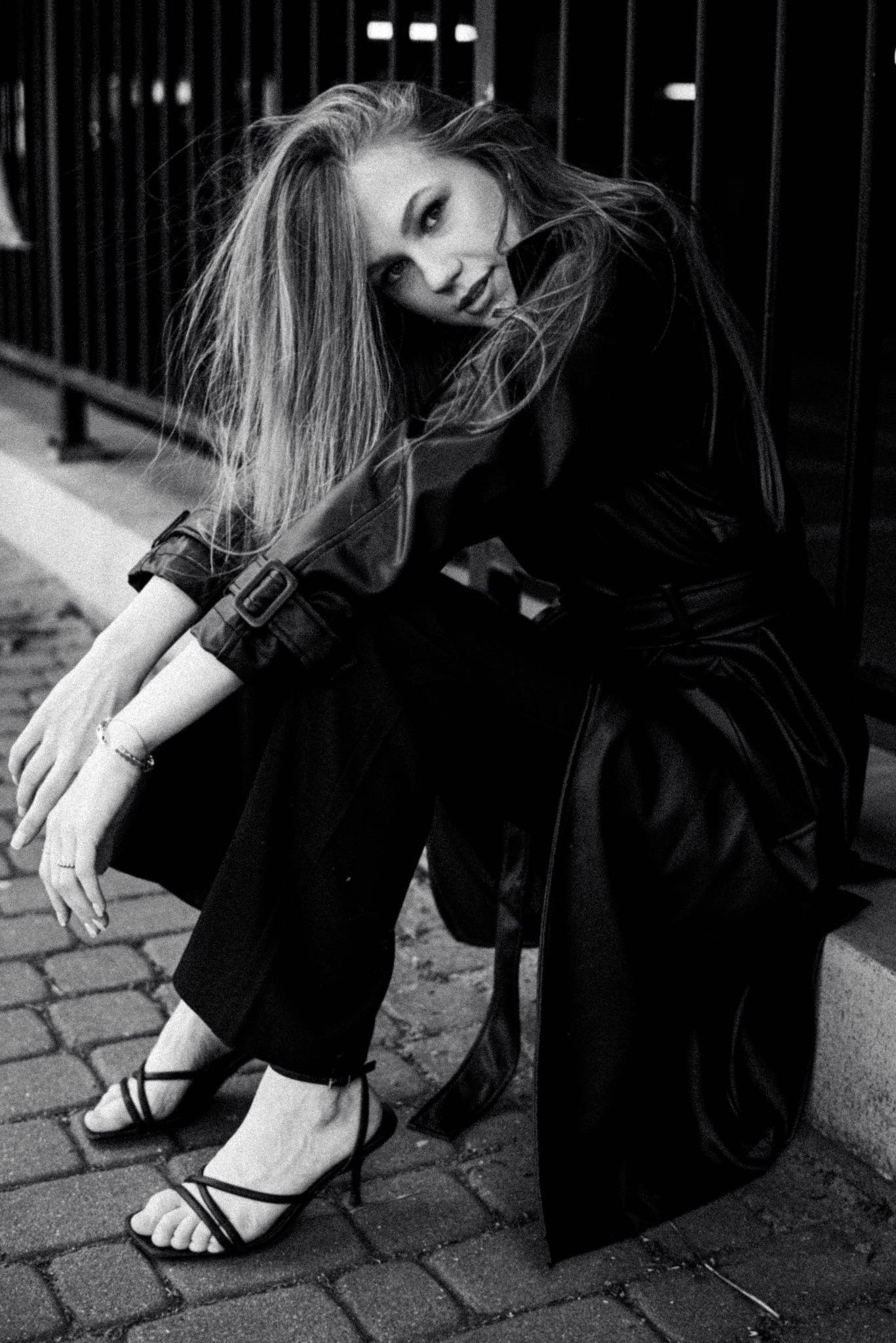 Czarno-białe zdjęcie kobiety w czarnym płaszczu i spodniach siedzącej na krawężniku przy metalowym ogrodzeniu, z delikatnym uśmiechem i spojrzeniem w obiektyw, włosy opadają na ramiona.