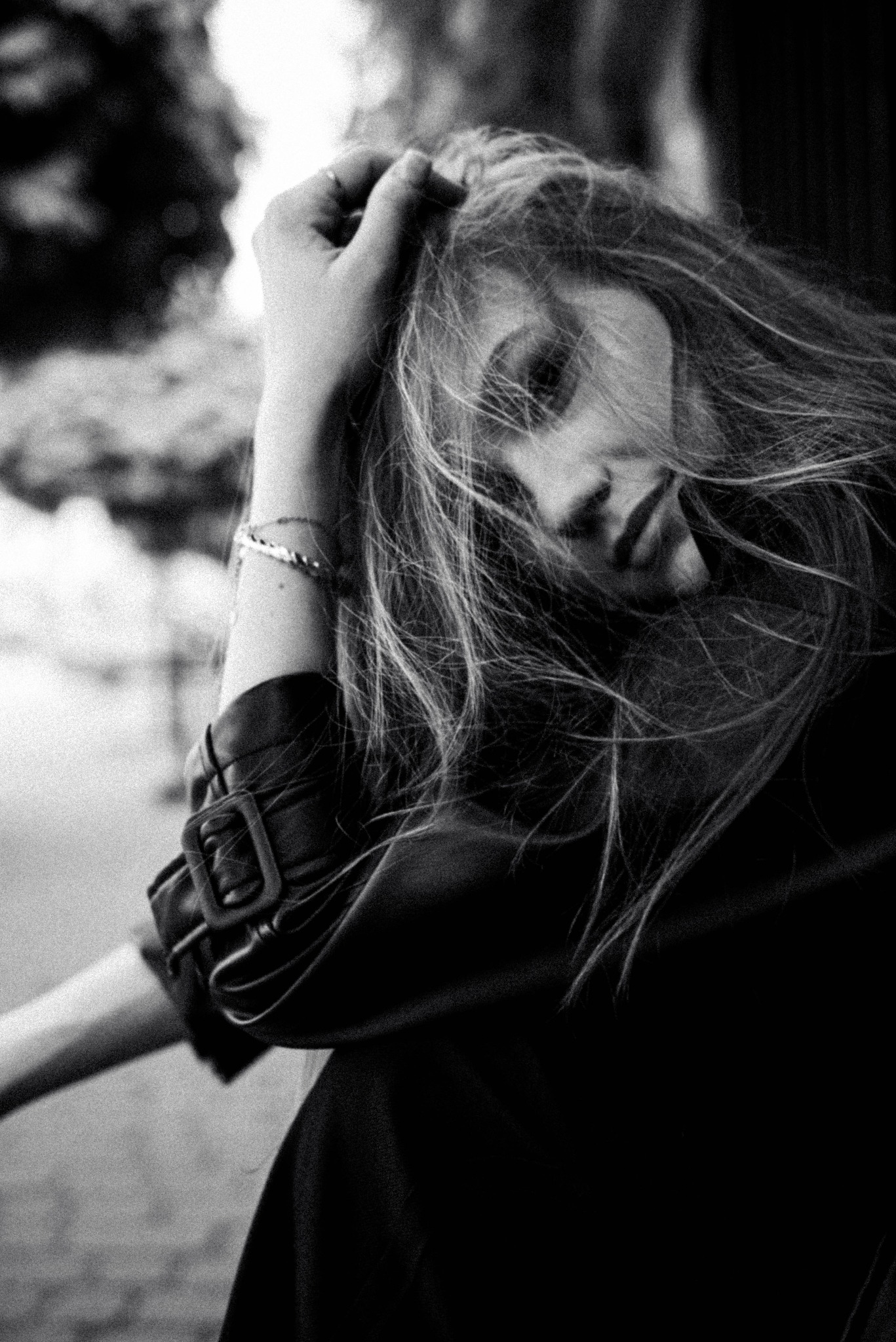 Czarno-białe portretowe ujęcie kobiety z długimi włosami, opierającej głowę na dłoni, ubranej w skórzaną kurtkę z widoczną klamrą, z zamyślonym wyrazem twarzy.