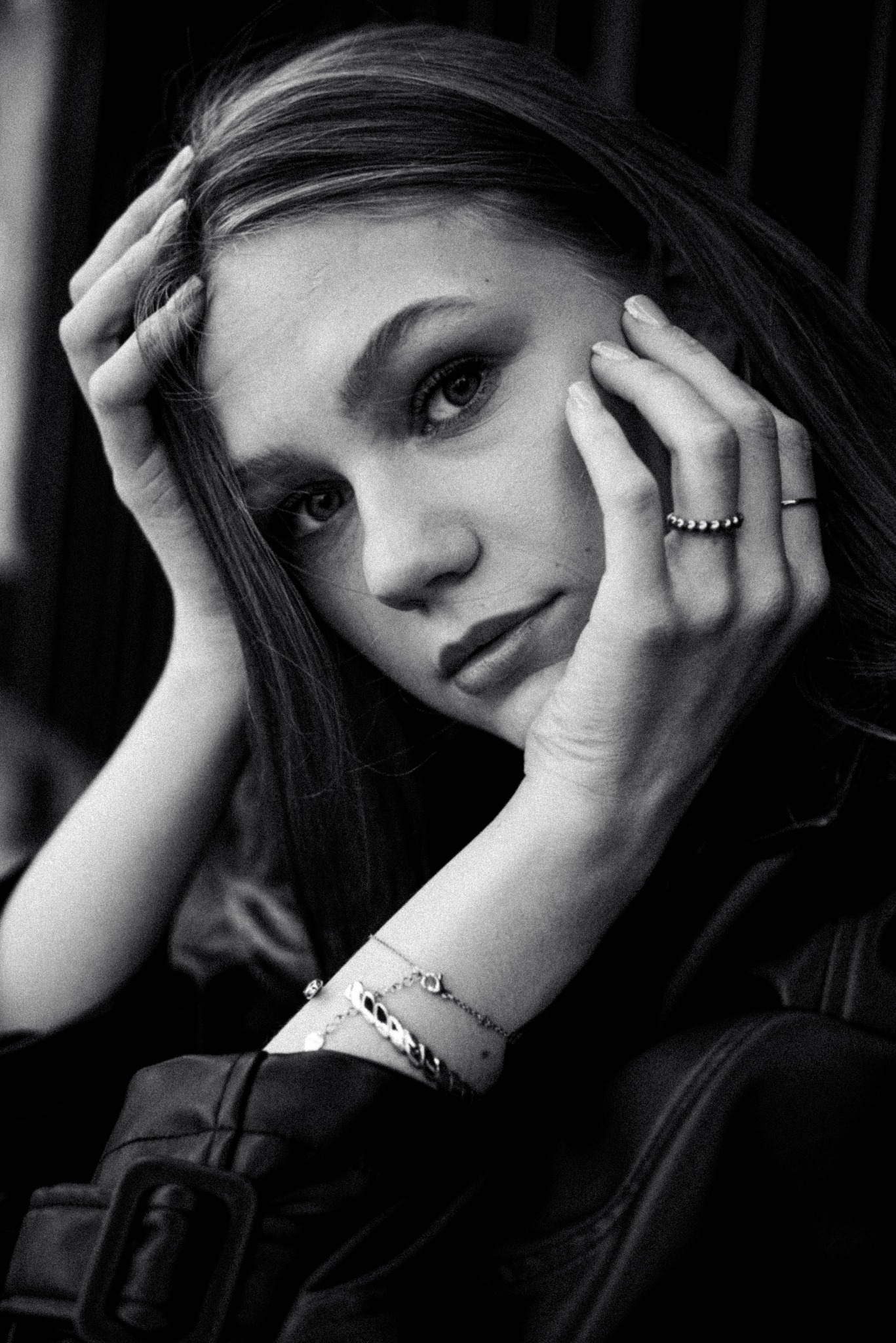 Portret młodej kobiety w czerni, z rękami uniesionymi do twarzy, ukazujący detale biżuterii: pierścionki i bransoletki, w monochromatycznej stylistyce.