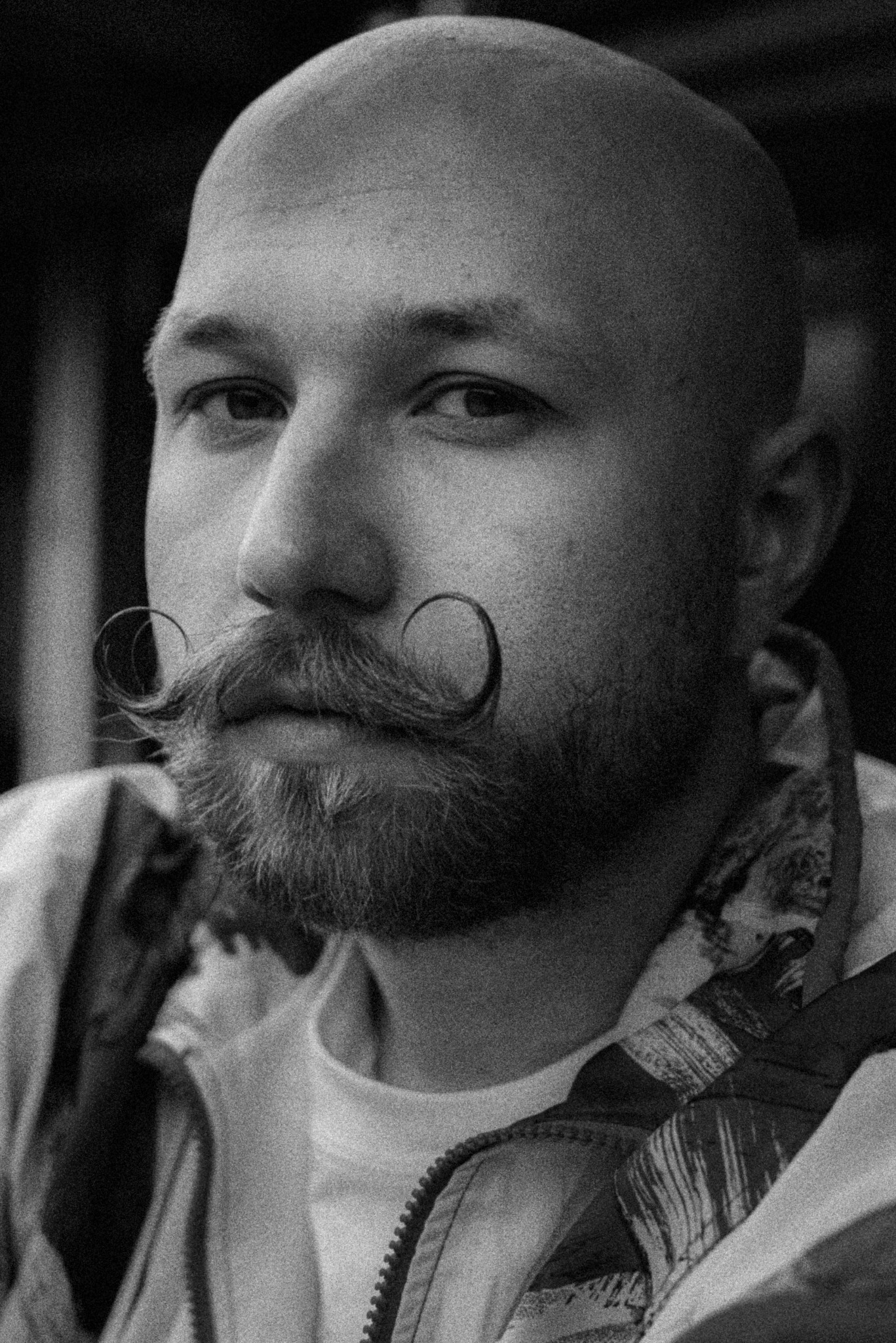 Czarno-białe portretowe ujęcie mężczyzny z ogoloną głową i charakterystycznymi, zakręconymi wąsami, ubrany w bluzę z zamkiem błyskawicznym.