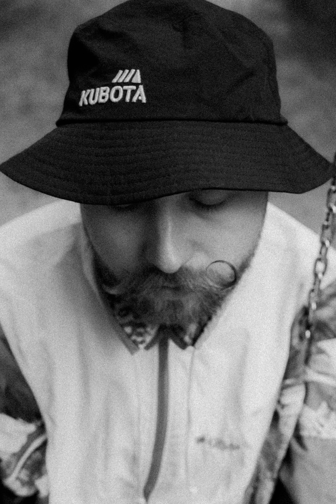 Portret mężczyzny z brodą i wąsami w czarnym kapeluszu z logo Kubota, czarno-białe zdjęcie z bliska.