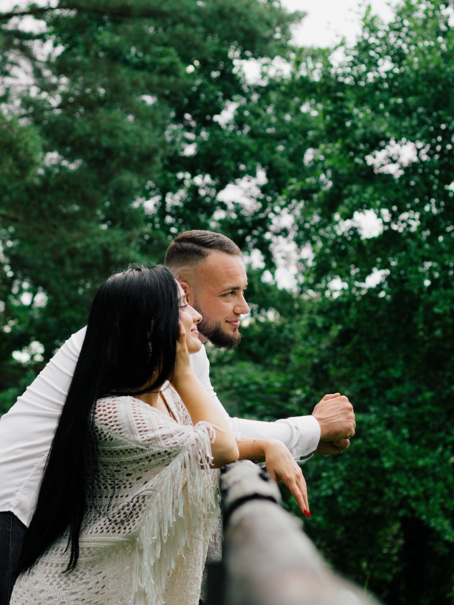 Para opiera się o drewnianą barierkę na tle zieleni; kobieta w ażurowym ponczo z frędzlami, mężczyzna w białej koszuli, oboje patrzą w dal.