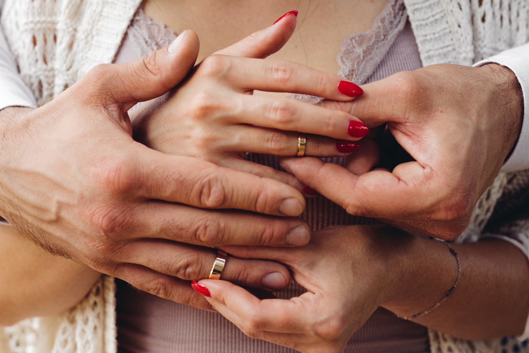 Zbliżenie dłoni pary z obrączkami ślubnymi; kobiece dłonie z czerwonym manicure trzymane w męskich dłoniach, delikatna bransoletka na nadgarstku kobiety, ażurowy sweter w tle.