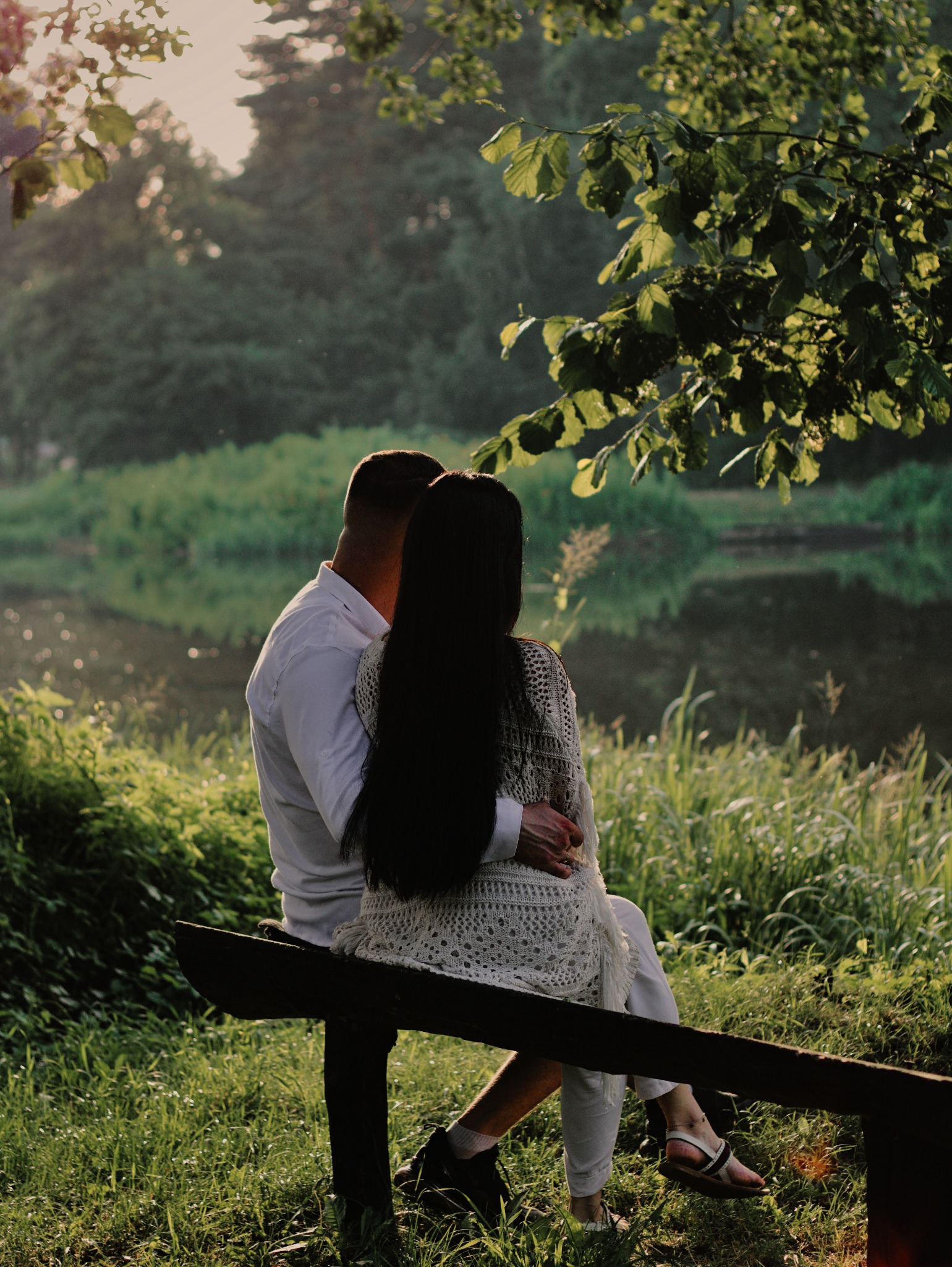 Para siedzi na drewnianej barierce nad brzegiem rzeki, obejmując się; ona ma długie, czarne włosy i ażurowy sweter, on w białej koszuli, w tle zieleń drzew i odbicie w wodzie.