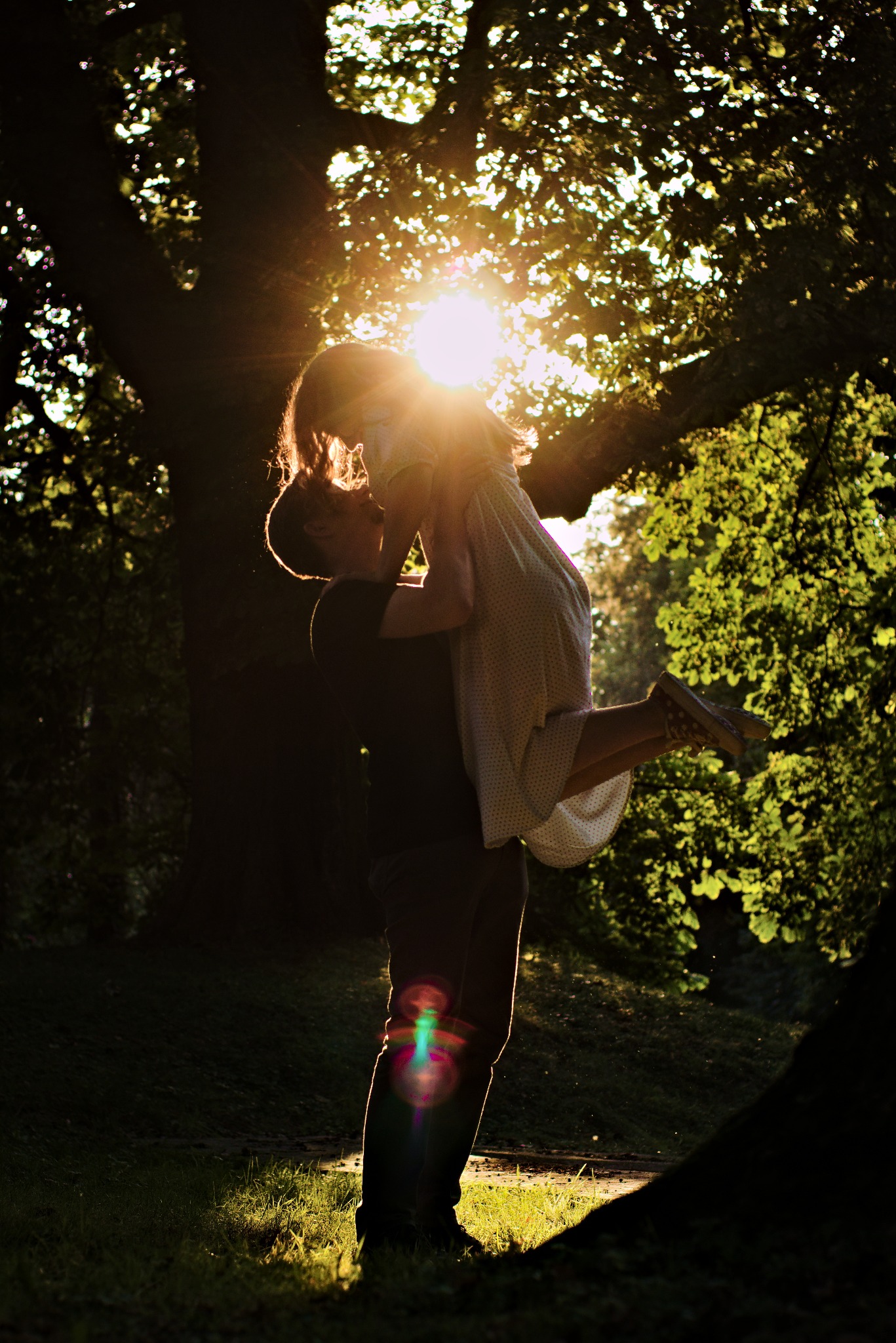 Zakochana para w silnym kontraście światła słonecznego: mężczyzna trzyma uniesioną kobietę na tle drzew z promieniami słońca przebijającymi przez liście.