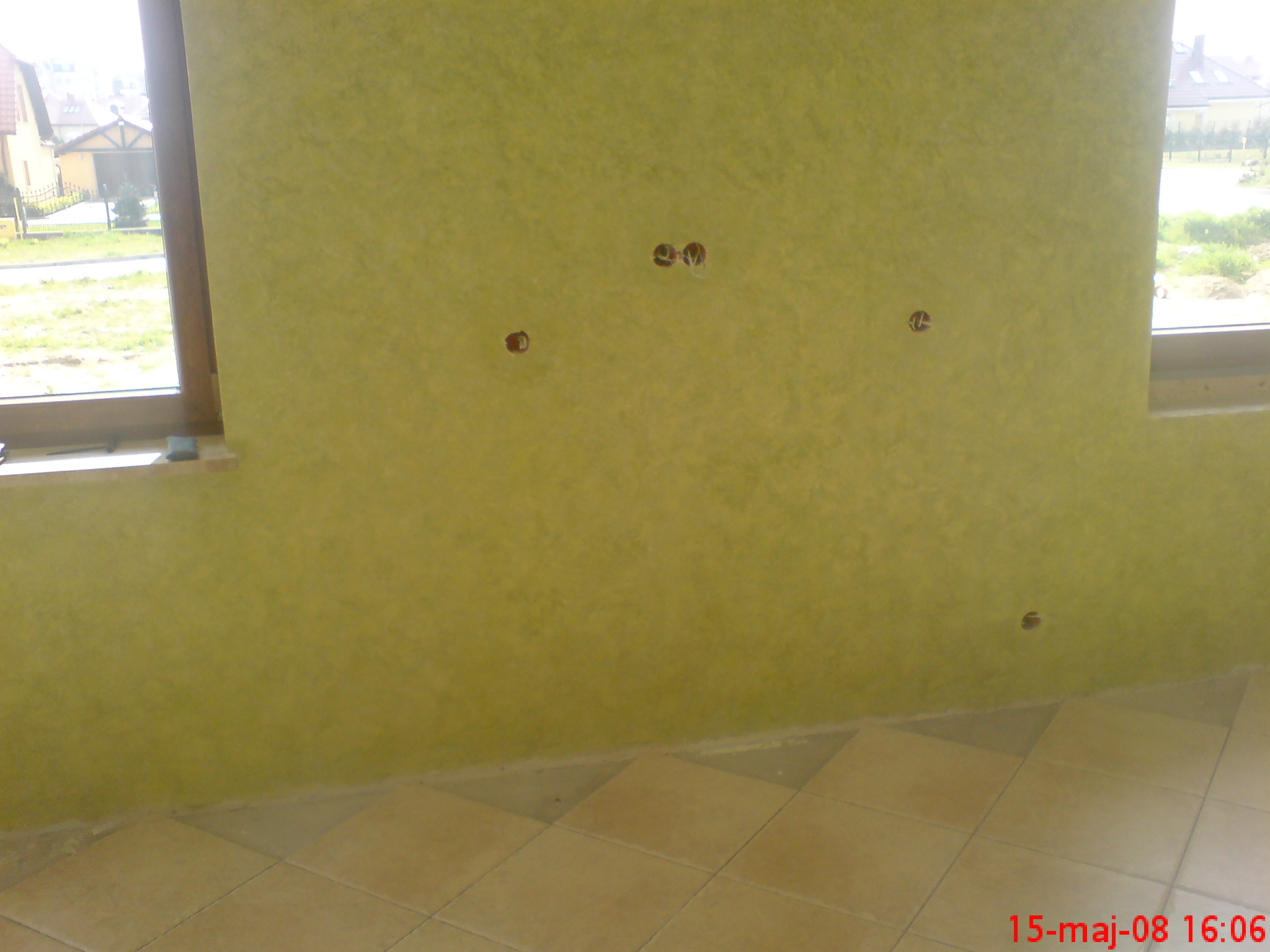 Żółta ściana z dekoracyjnym tynkiem akrylowym, widoczne otwory na gniazdka elektryczne, dolna część ściany przy podłodze wykończona fugą, w tle okno z widokiem na dom i zieleń.