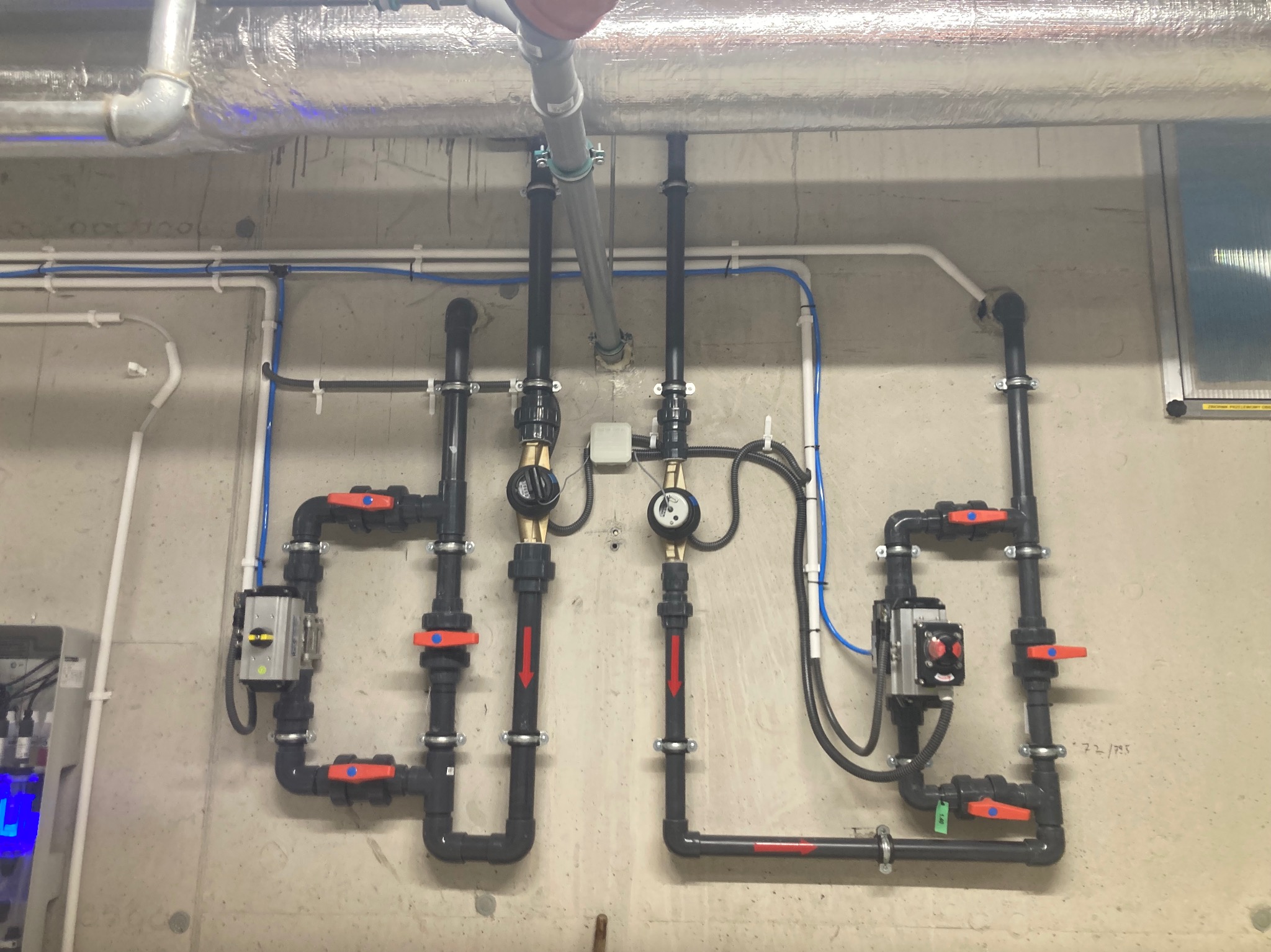 Instalacja hydrauliczna z czarnych rur i czerwonymi zaworami na szarej ścianie, widoczne liczniki i skrzynka sterująca, instalacja pod sufitem.