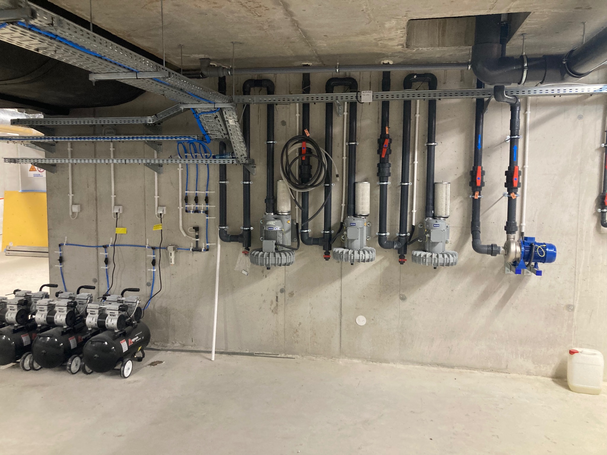 Pomieszczenie techniczne z rzędami pomp i systemem rur pod sufitem, widoczne również kompresory na kółkach i kanistry