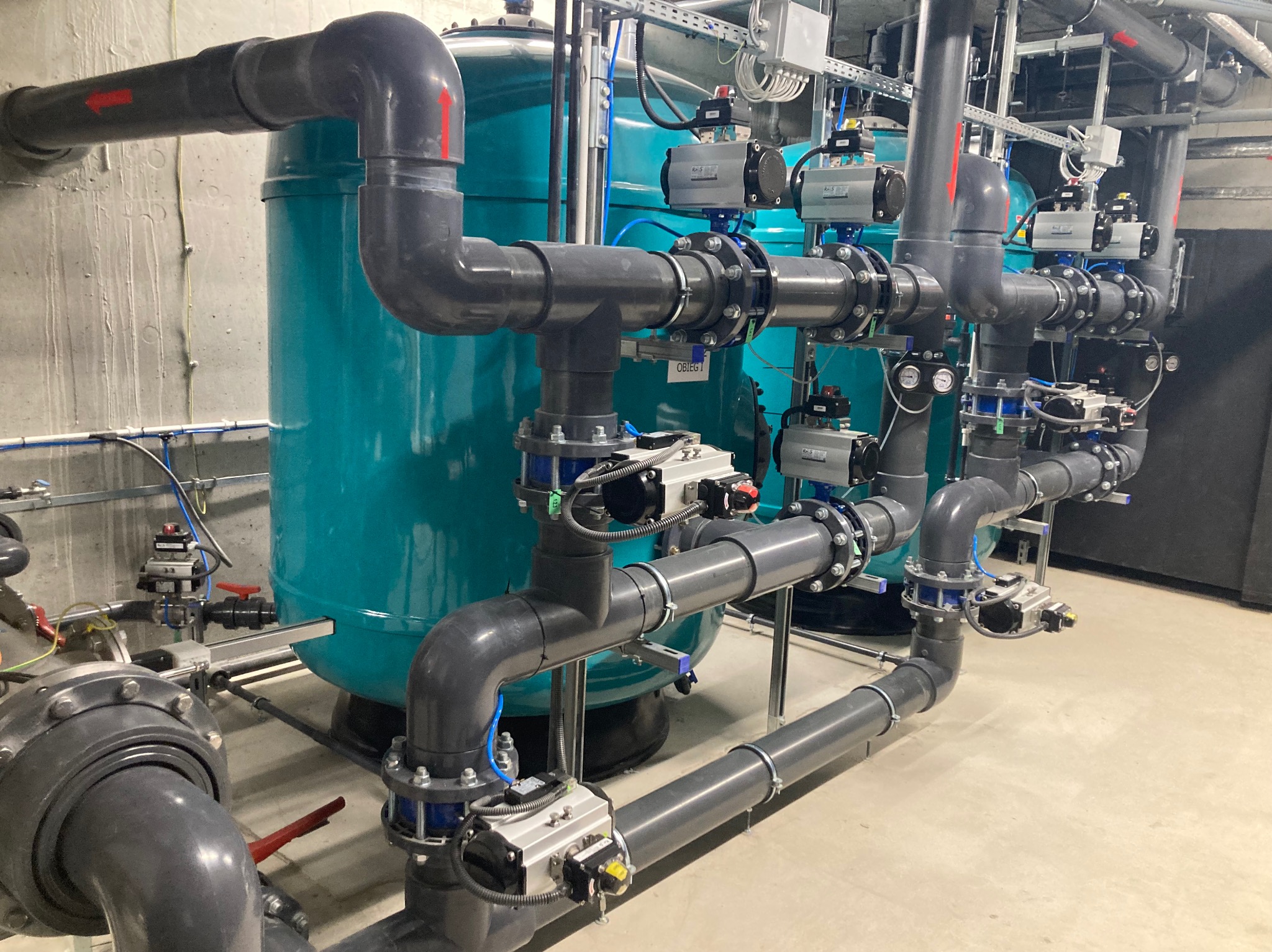 Instalacja filtracyjna z rurami PCV i zbiornikami ciśnieniowymi, widoczne liczne zawory i manometry, całość w pomieszczeniu technicznym.
