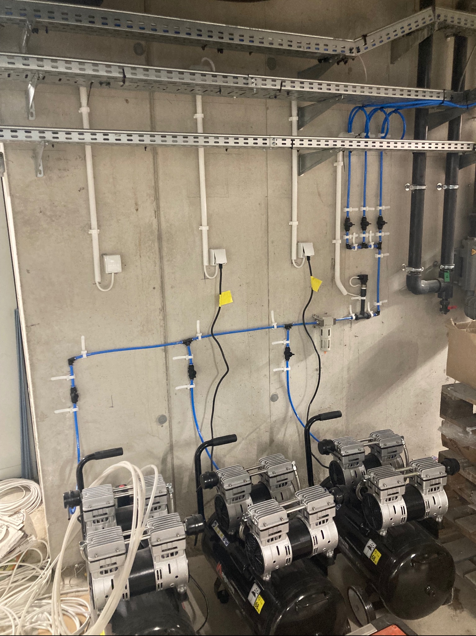 Sprężarki powietrza podłączone do instalacji pneumatycznej na tle surowej betonowej ściany z widocznymi rurami i kablami w metalowych korytach