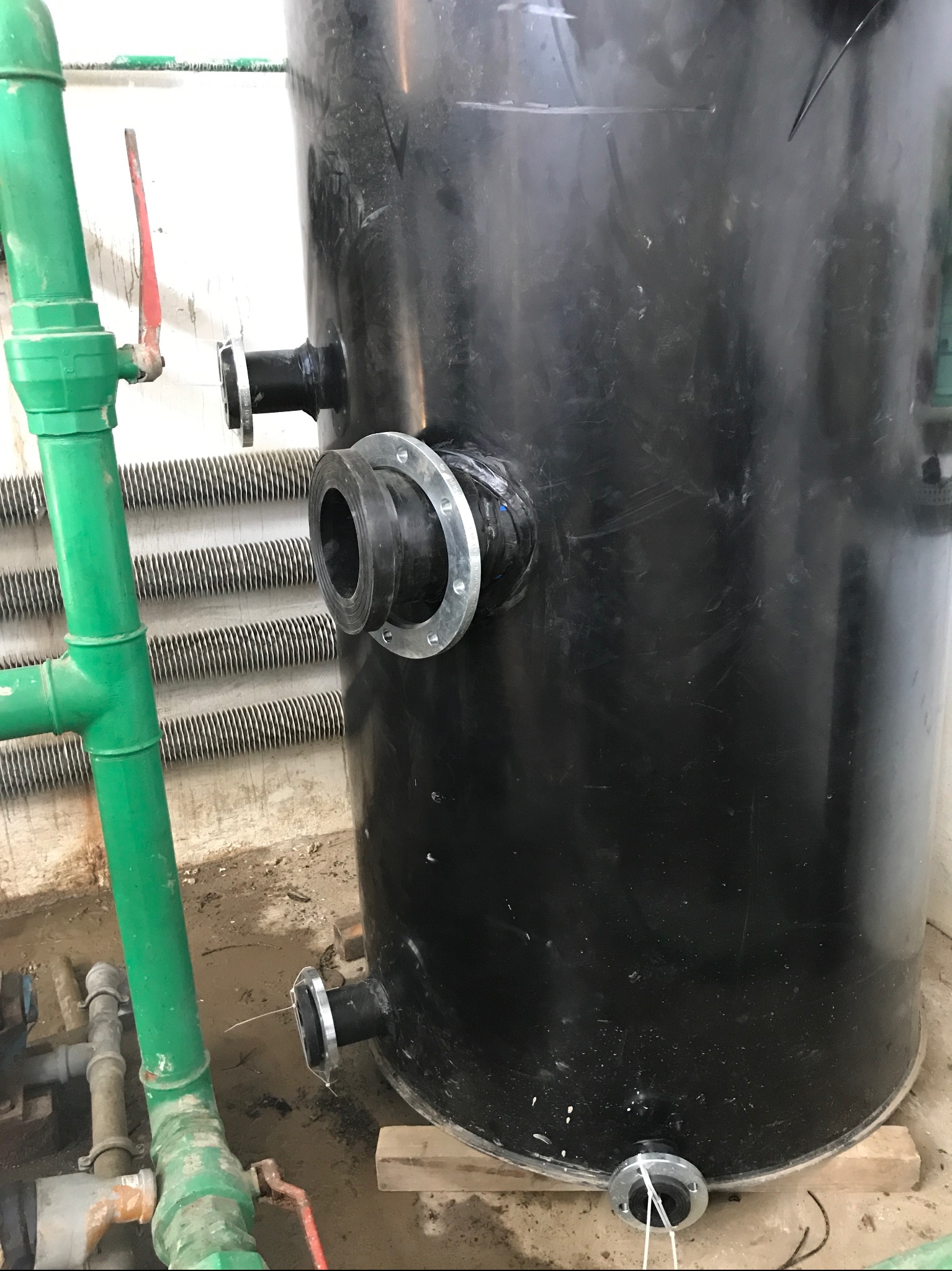 Czarny zbiornik z podłączeniami rurowymi i kołnierzami, stojący na drewnianych podkładkach, obok zielonej instalacji rurowej z zaworem.