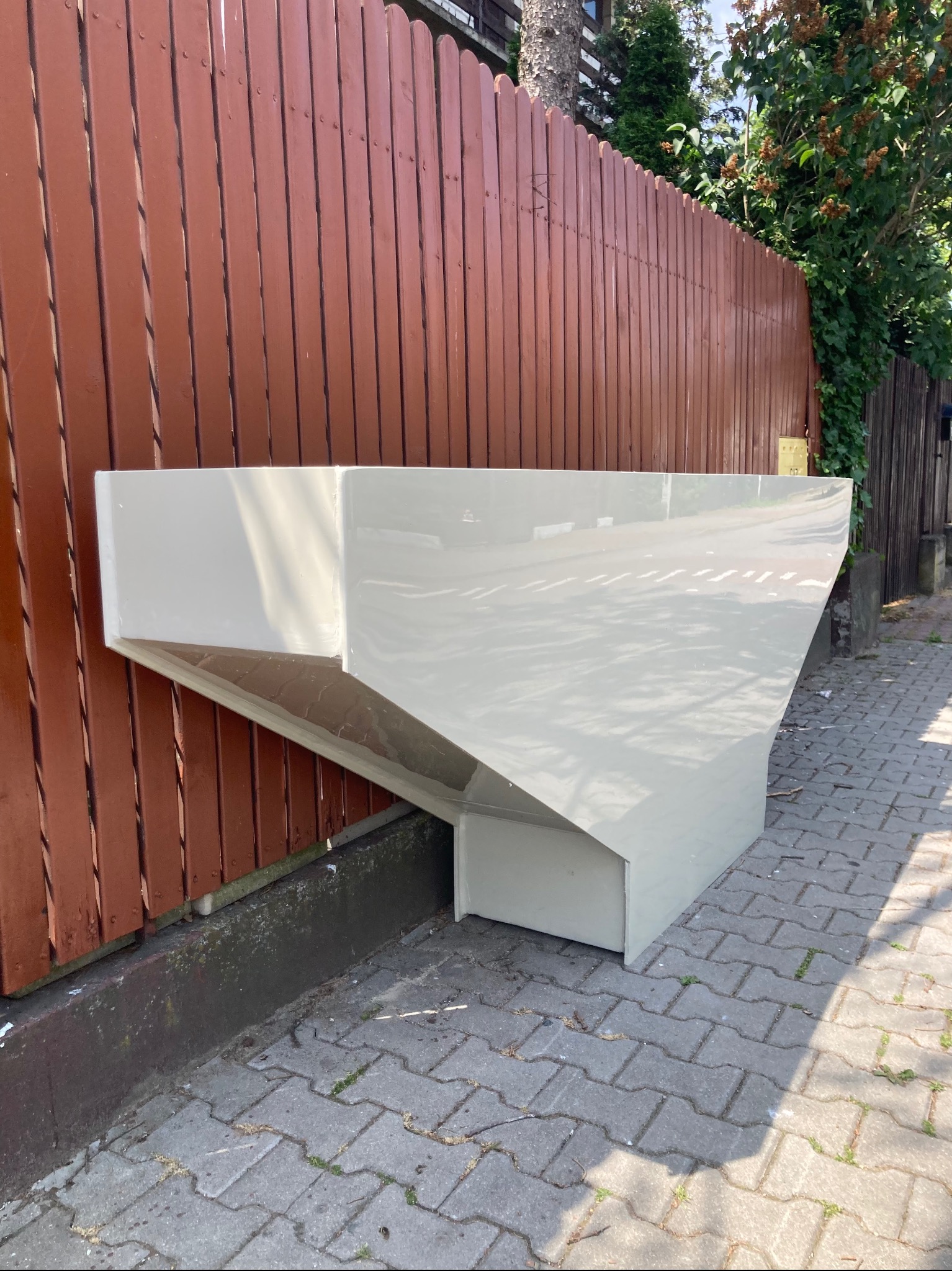 Prefabrykowana, biała konstrukcja z tworzywa sztucznego, prawdopodobnie fragment większego zbiornika, oparta o brązowy płot z desek na brukowanym chodniku.