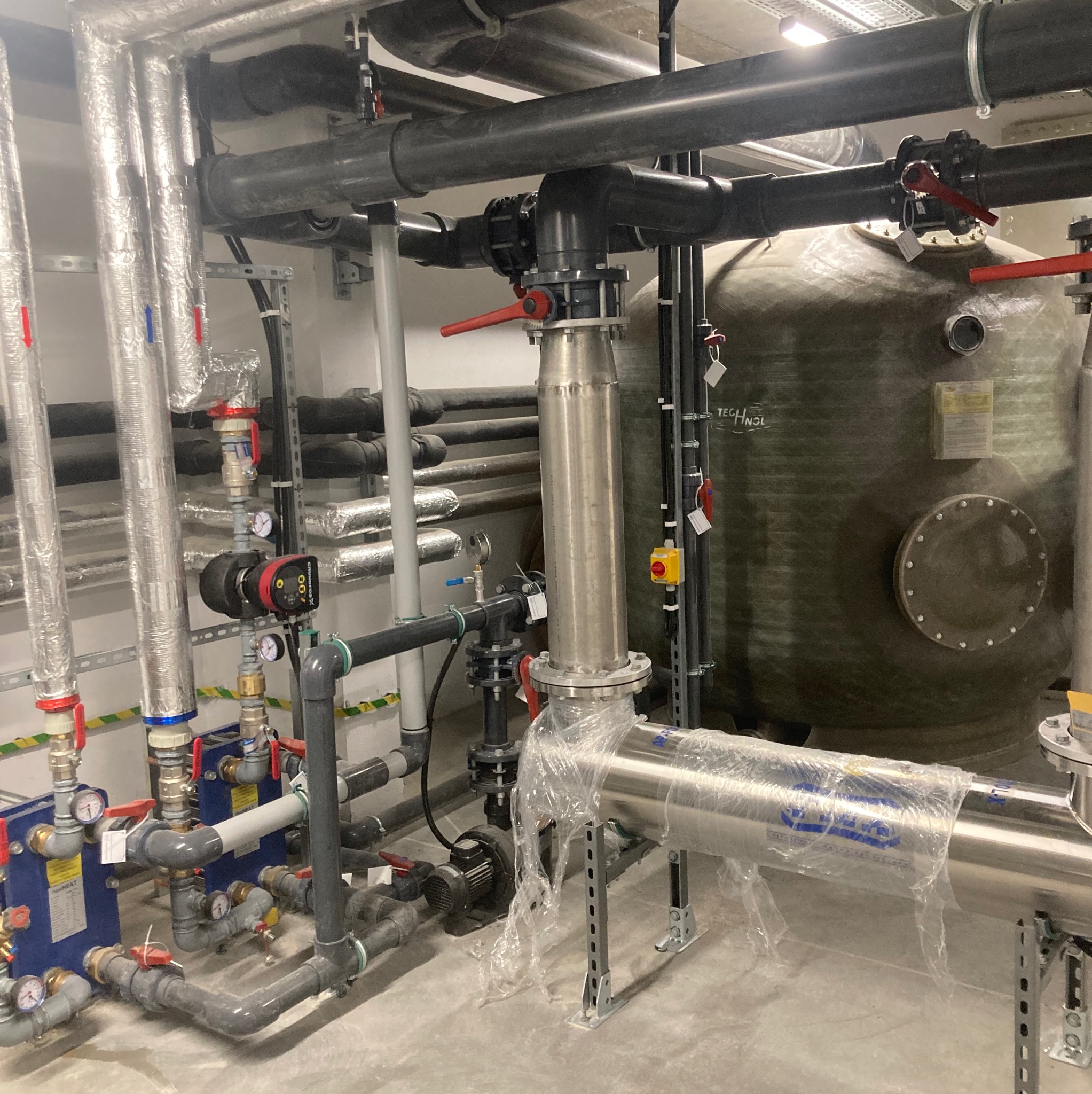 Kompleksowy system filtracji wody z rurami PCV, stalowymi elementami, zaworami z czerwonymi uchwytami, manometrami i dużym zbiornikiem filtracyjnym z włókna szklanego w pomieszczeniu technicznym.