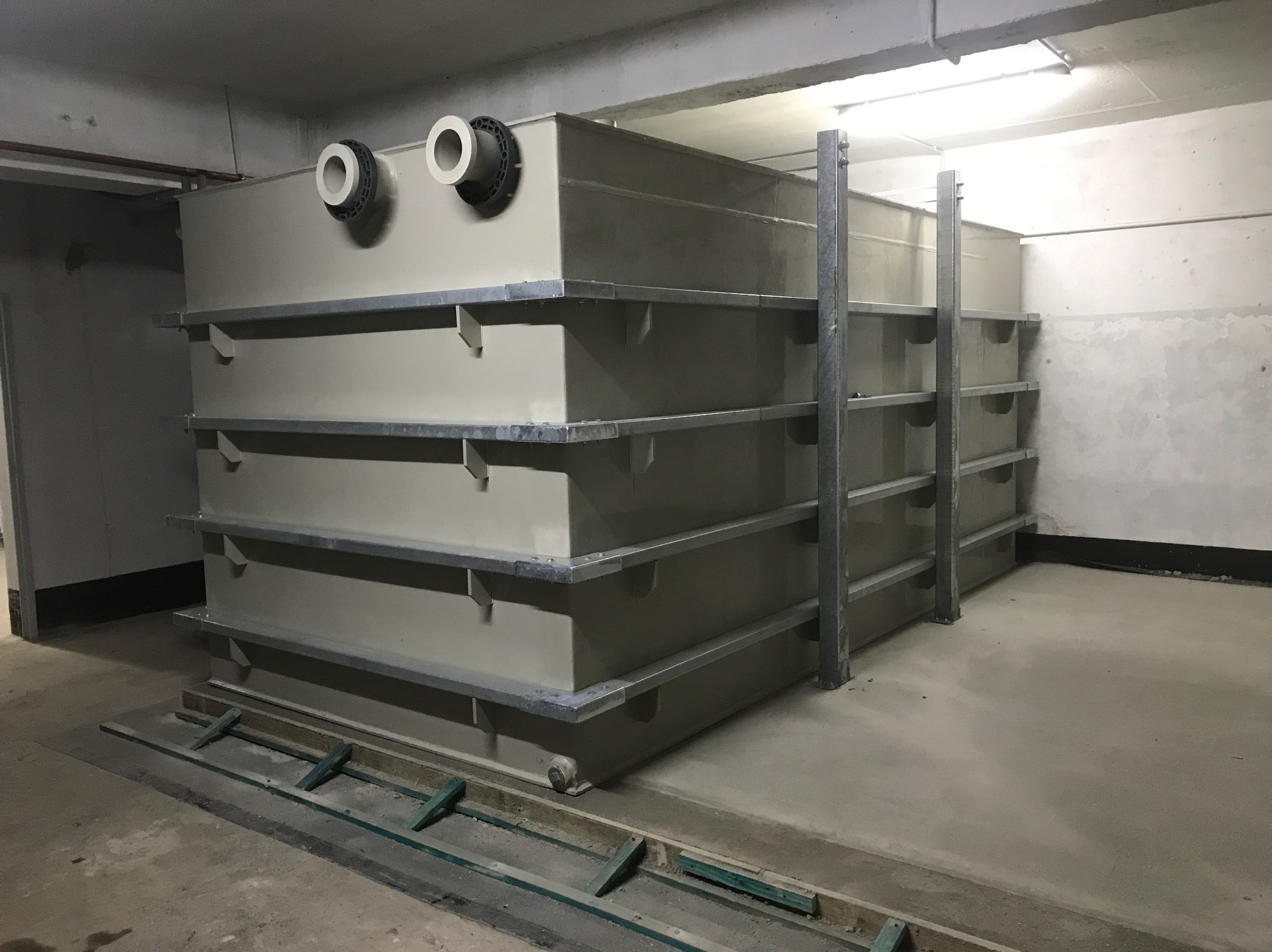 Duży, beżowy zbiornik z rurami i metalowymi wspornikami w szarym pomieszczeniu, prawdopodobnie w trakcie instalacji.