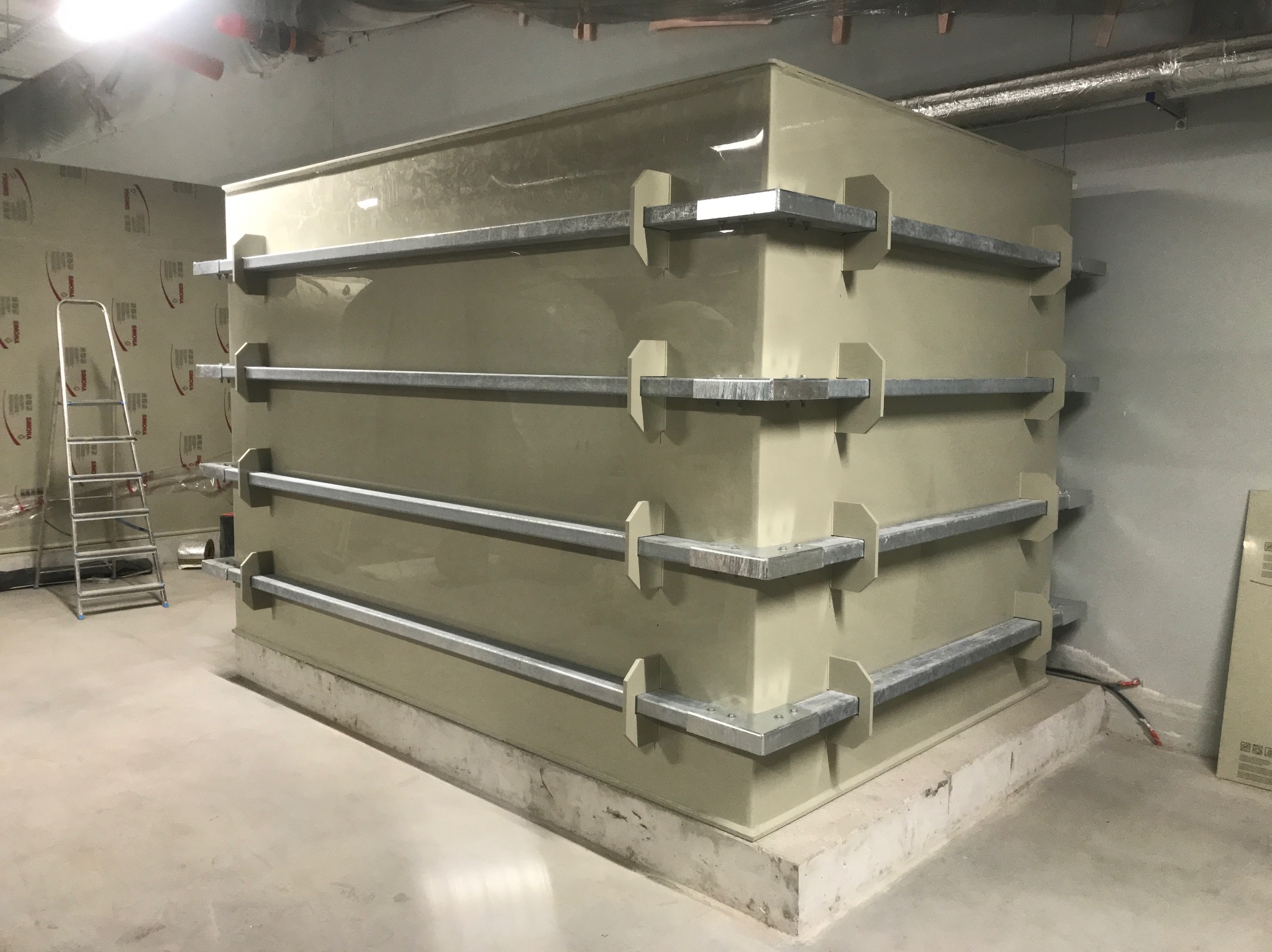 Duży, beżowy zbiornik z metalowymi wzmocnieniami w surowym wnętrzu budynku, obok drabina aluminiowa, widoczne rury pod sufitem.