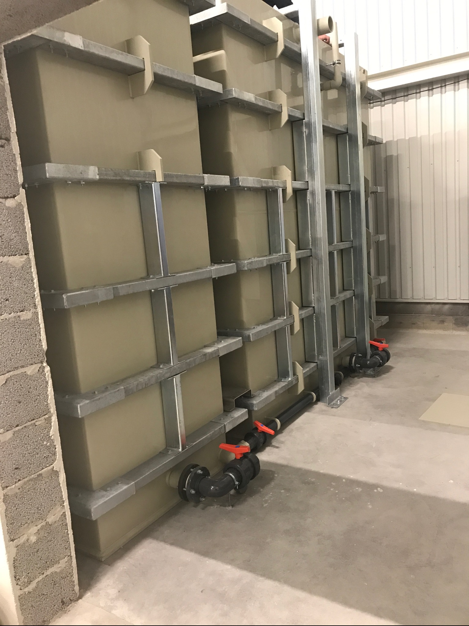 Rząd dużych, beżowych zbiorników z metalowymi ramami wewnątrz budynku, z widocznymi rurami i zaworami na dole, przy ścianie z betonowych pustaków.