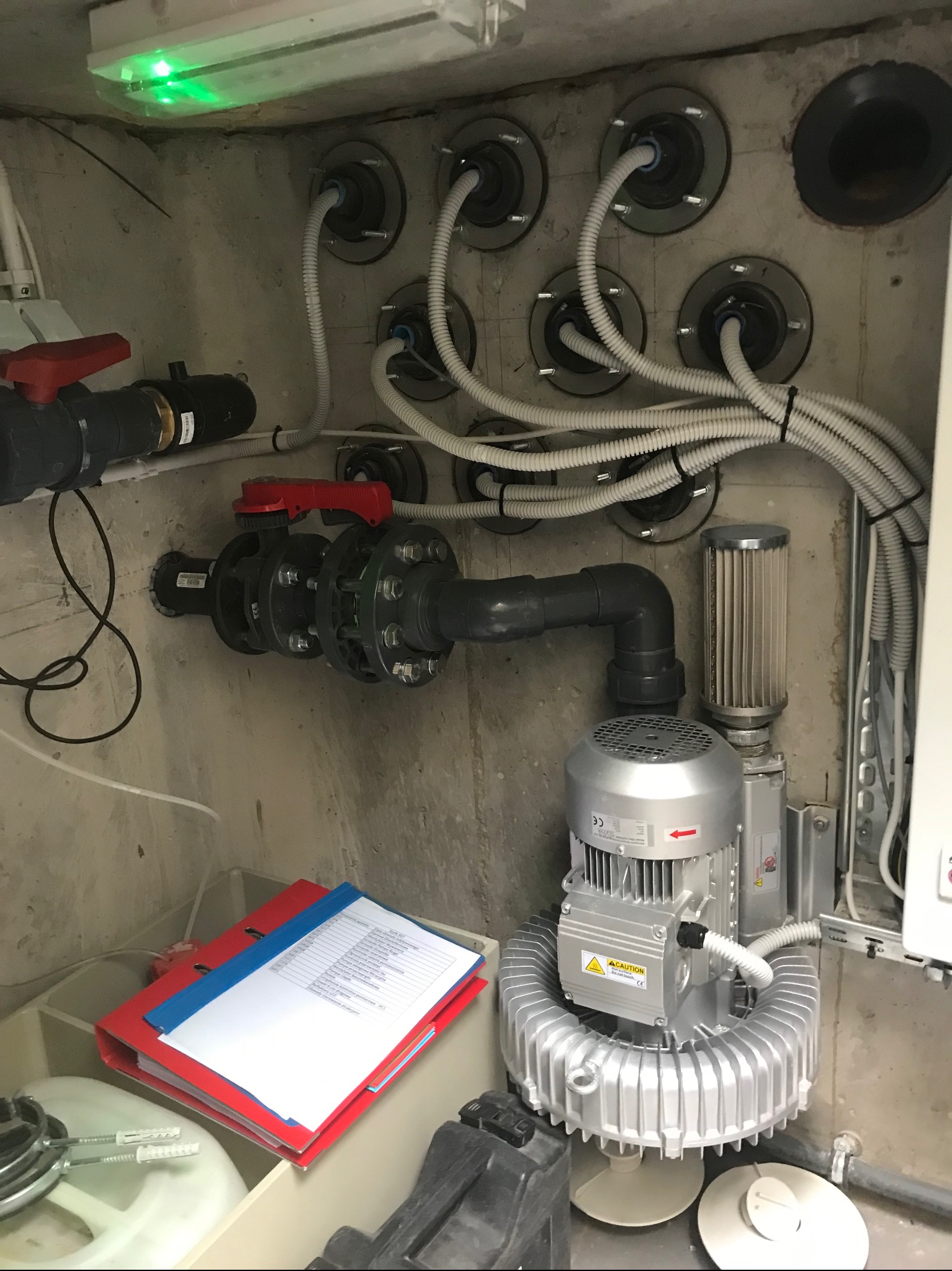 Pomieszczenie techniczne z instalacją filtracyjną i pompą do cyrkulacji wody, widoczne rury, zawory, silnik oraz dokumentacja w segregatorach.