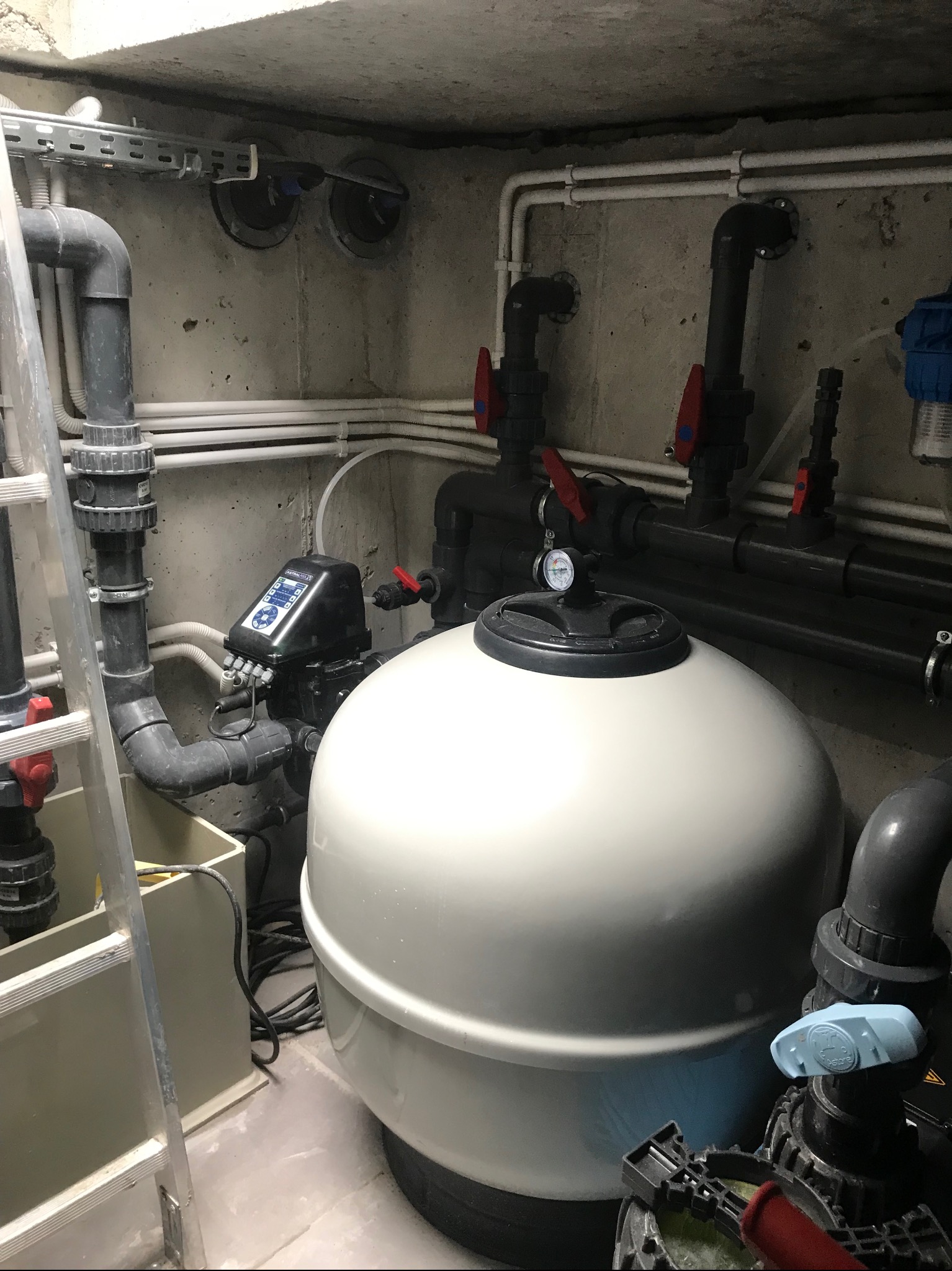 Pomieszczenie techniczne z dużym filtrem piaskowym do basenu, rurami PCV, zaworami i drabinką aluminiową w tle.