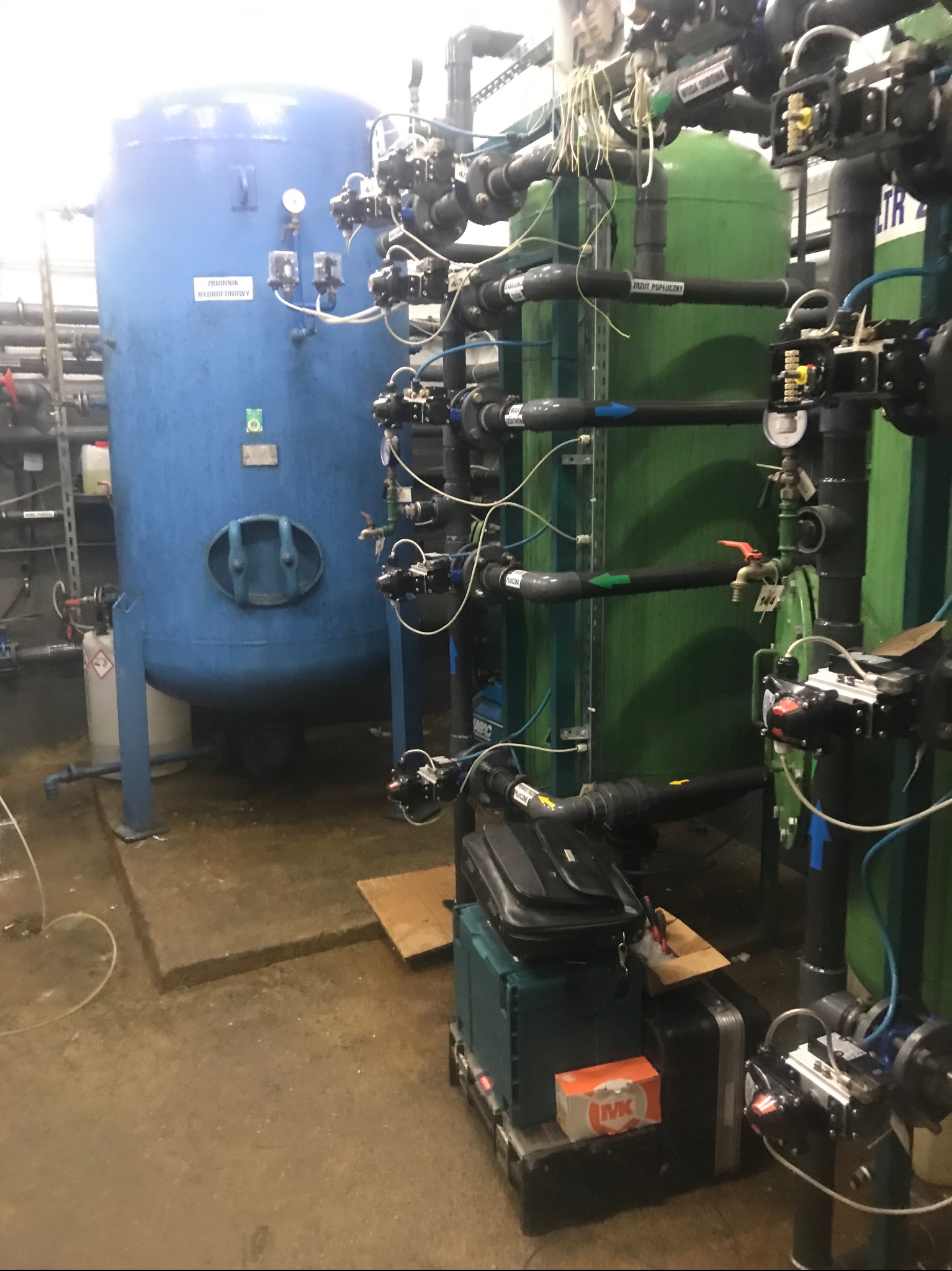 Instalacja filtracyjna z dwoma zbiornikami, niebieskim i zielonym, oraz systemem rur i zaworów; na podłodze skrzynki narzędziowe i walizki.