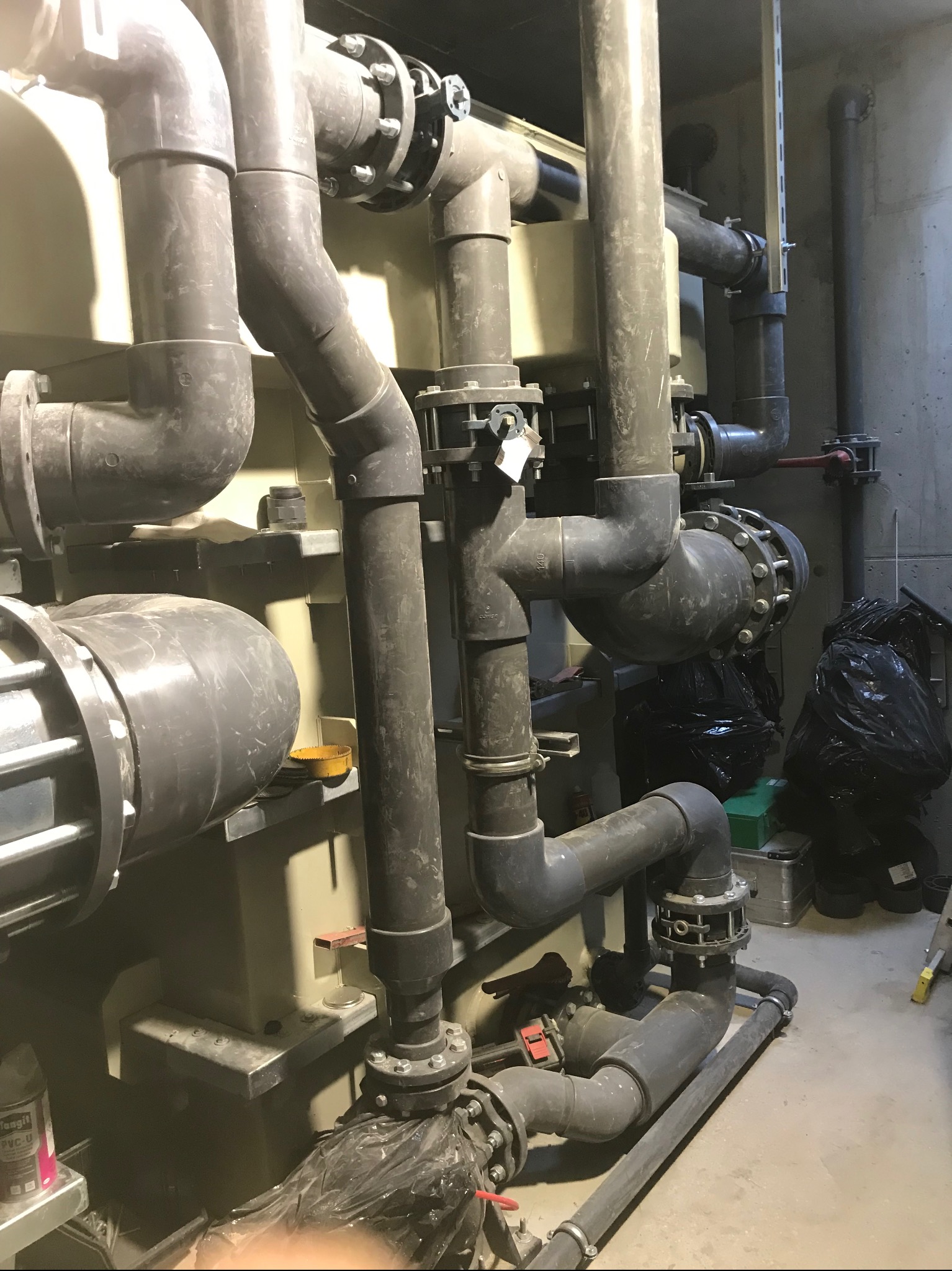 Instalacja rurowa z zaworami i filtrami, prawdopodobnie w pomieszczeniu technicznym basenu, widoczne czarne worki na śmieci i drabina.