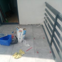 Zbigniew Niewiadomy - Szare płytki podłogowe na balkonie w trakcie układania, widoczny worek z klejem, wiadro, poziomica i plastikowe kliny dystansowe.