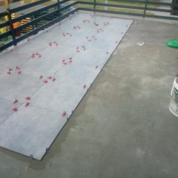 Zbigniew Niewiadomy - Szare płytki ułożone na balkonie, z czerwonymi klipsami poziomującymi, widoczne wiadro z zaprawą i narzędzia.