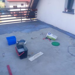 Zbigniew Niewiadomy - Hydroizolacja balkonu w Otmuchowie w trakcie realizacji: widoczne wiadra z materiałem, paczka z zaprawą, narzędzia i częściowo pokryta podłoga balkonu. Balustrada metalowa w tle.