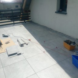 Zbigniew Niewiadomy - Układanie szarych płytek na balkonie z narzędziami: przecinarka, poziomica, szpachelka, pojemniki z zaprawą, czerwony sznur traserski.