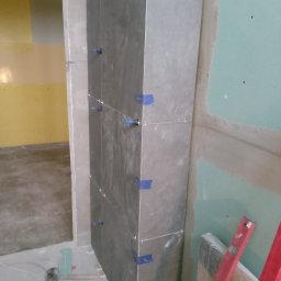 Zbigniew Niewiadomy - Fragment ściany obłożonej ciemnymi płytkami, zabezpieczonymi niebieską taśmą i krzyżykami dystansowymi, obok ściana z płyty gipsowo-kartonowej, na podłodze narzędzia i resztki płytek.