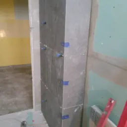 Fragment ściany obłożonej ciemnymi płytkami, zabezpieczonymi niebieską taśmą i krzyżykami dystansowymi, obok ściana z płyty gipsowo-kartonowej, na podłodze narzędzia i resztki płytek.