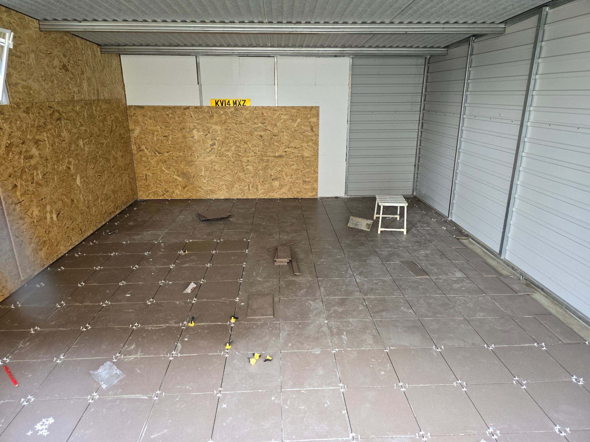 Montaż brązowych płytek podłogowych w garażu, widoczne krzyżyki dystansowe, narzędzia i fragment ściany z płyt OSB.