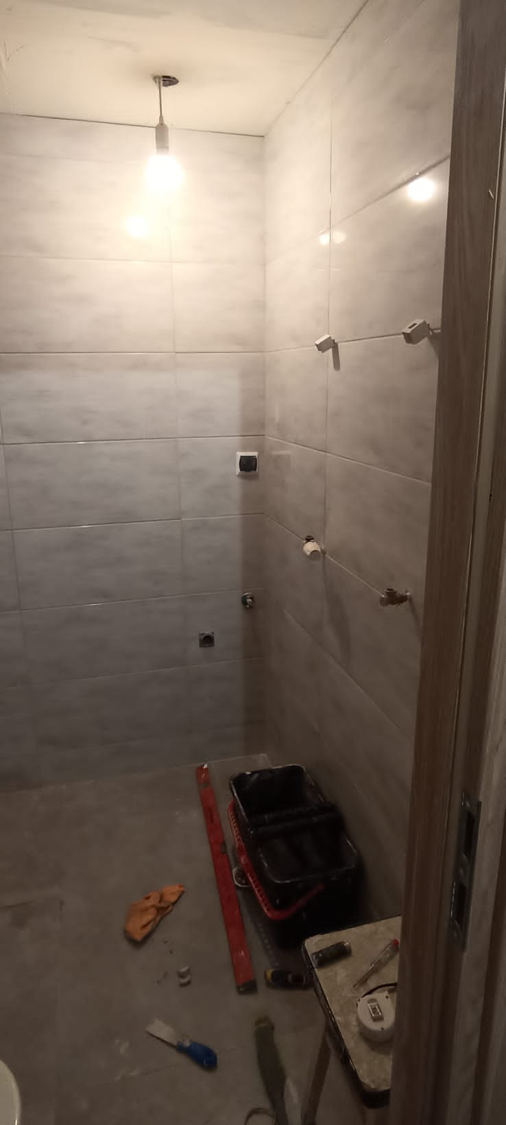 Łazienka w trakcie układania płytek, widoczne narzędzia: poziomica, wiadro, szpachelka, lampa sufitowa z żarówką, częściowo zamontowane akcesoria łazienkowe.