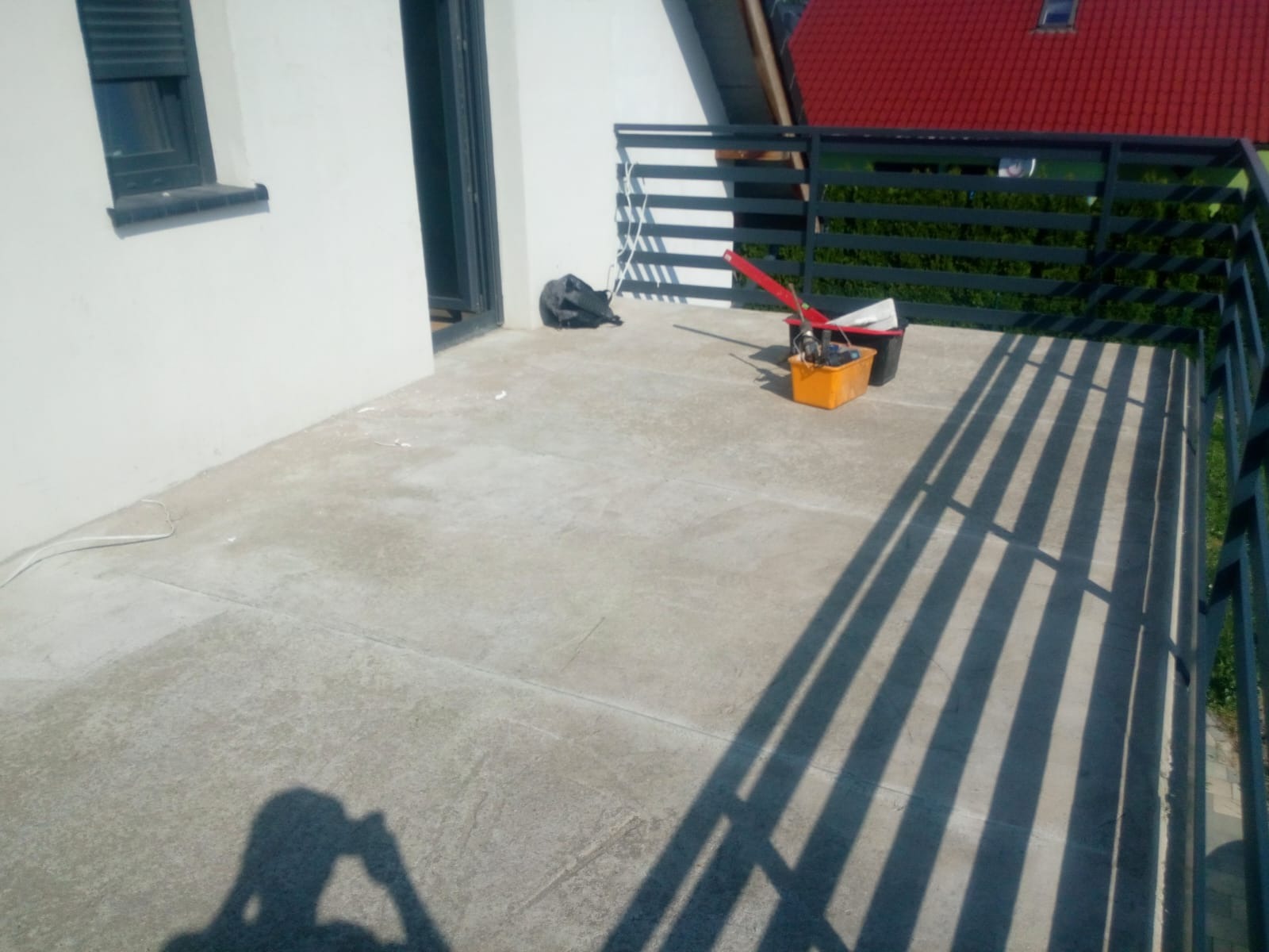 Balkon z szarą, betonową posadzką w trakcie przygotowań do malowania, widoczne narzędzia malarskie w pomarańczowym wiadrze i cień osoby robiącej zdjęcie.