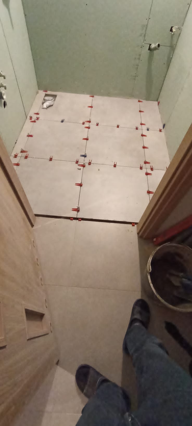 Łazienka w trakcie układania płytek podłogowych z użyciem poziomujących klipsów i klinów, widoczne niezabudowane rury i elementy instalacji wodnej, częściowo wykończone ściany z płyty...