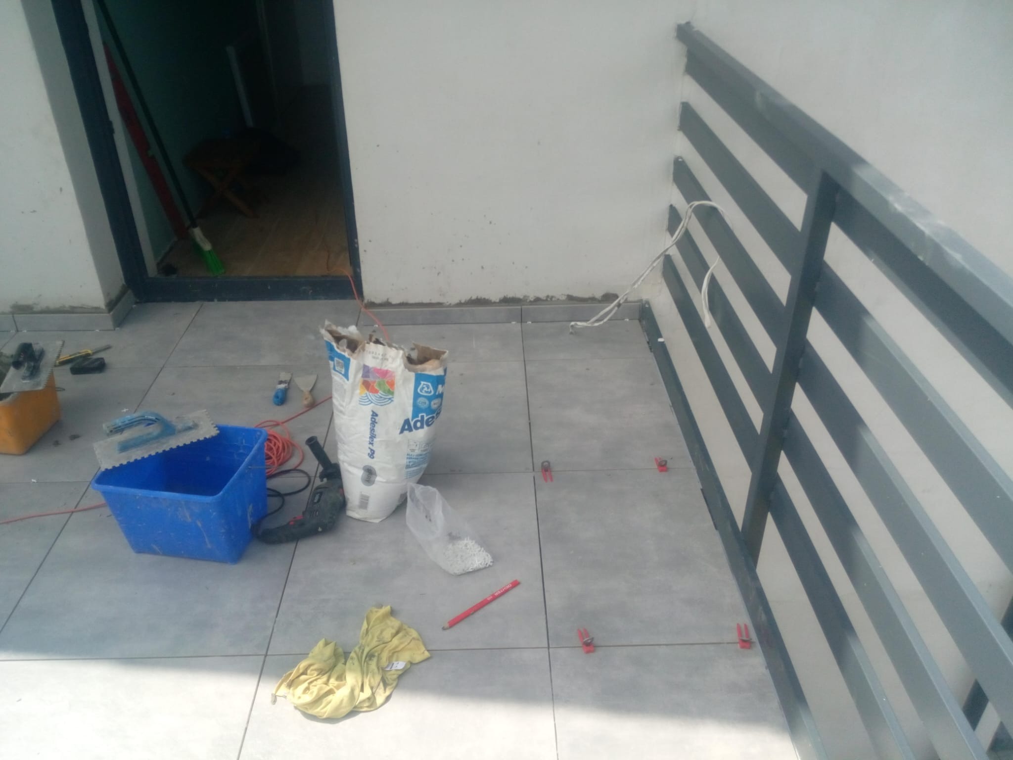 Szare płytki podłogowe na balkonie w trakcie układania, widoczny worek z klejem, wiadro, poziomica i plastikowe kliny dystansowe.