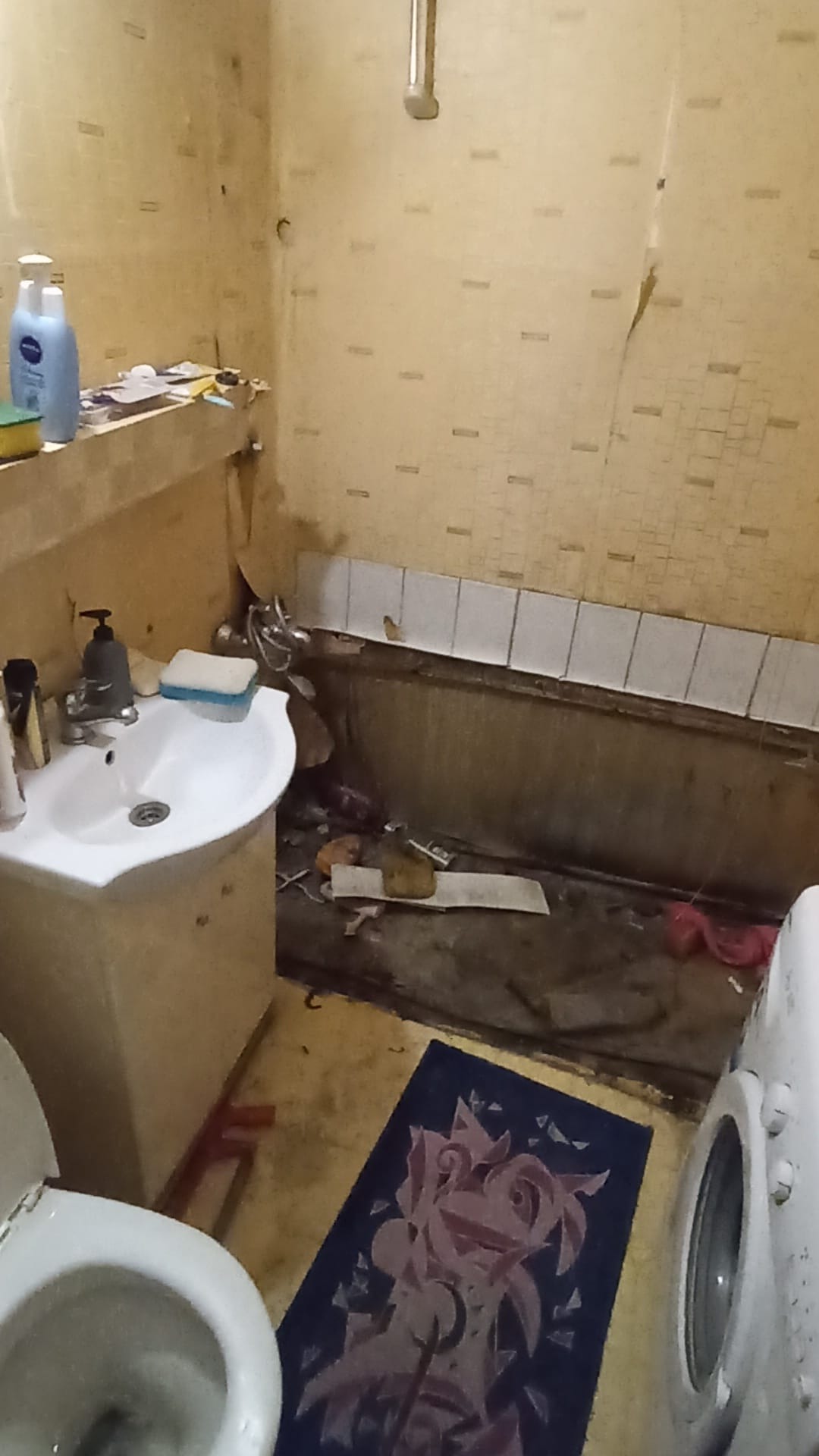 Zniszczona łazienka z odpadającą tapetą, brudną wanną, umywalką i toaletą, widoczny dywanik na podłodze i pralka z boku.