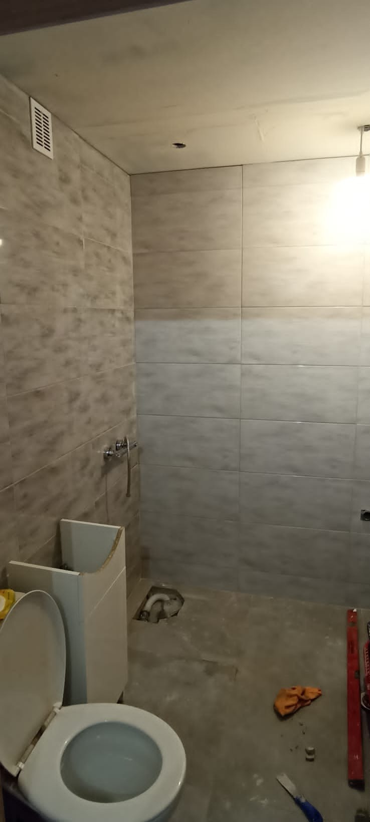Remont łazienki w trakcie: położone płytki w odcieniach szarości, widoczny otwór kanalizacyjny w podłodze, częściowo zamontowana toaleta i narzędzia budowlane.