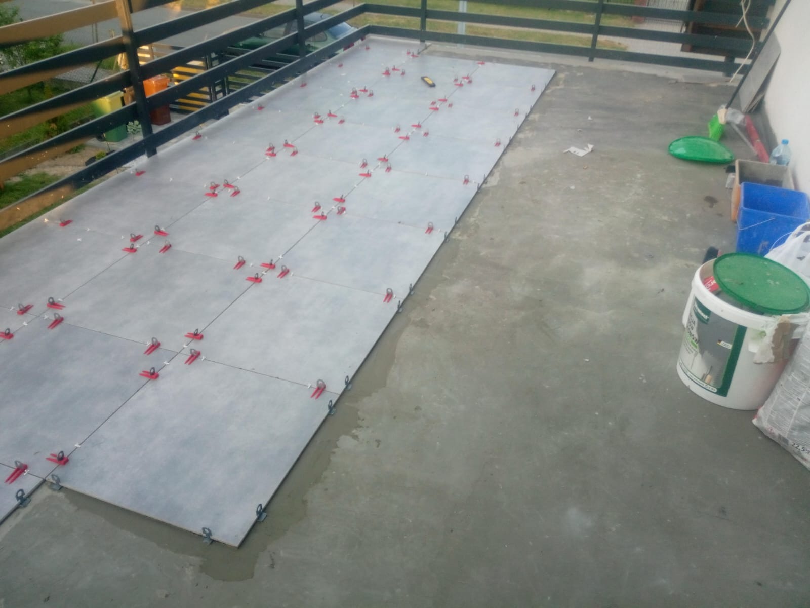 Szare płytki ułożone na balkonie, z czerwonymi klipsami poziomującymi, widoczne wiadro z zaprawą i narzędzia.