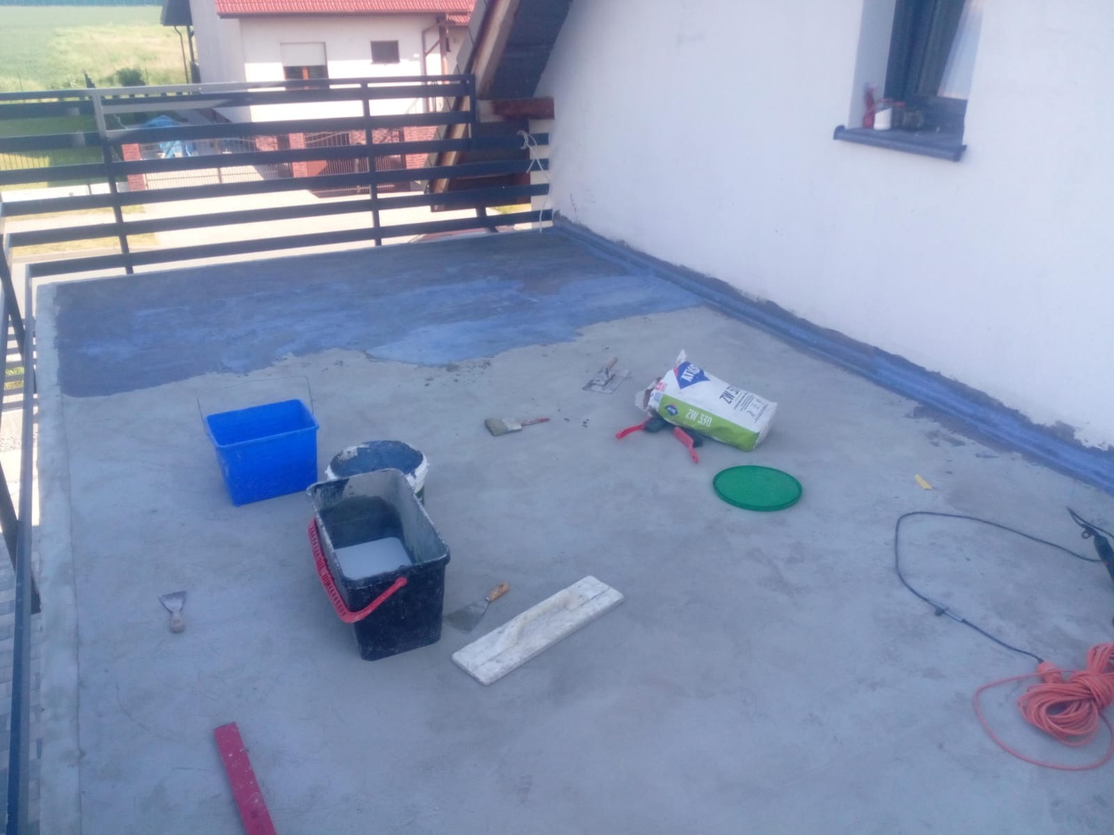Hydroizolacja balkonu w Otmuchowie w trakcie realizacji: widoczne wiadra z materiałem, paczka z zaprawą, narzędzia i częściowo pokryta podłoga balkonu. Balustrada metalowa w tle.
