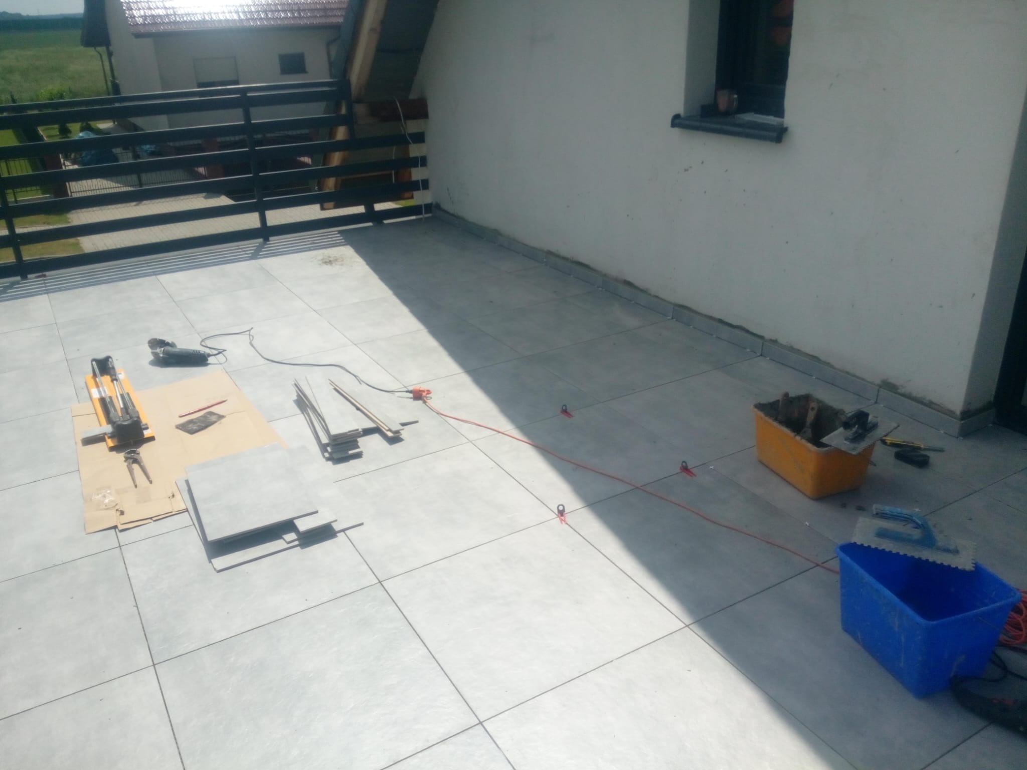 Układanie szarych płytek na balkonie z narzędziami: przecinarka, poziomica, szpachelka, pojemniki z zaprawą, czerwony sznur traserski.