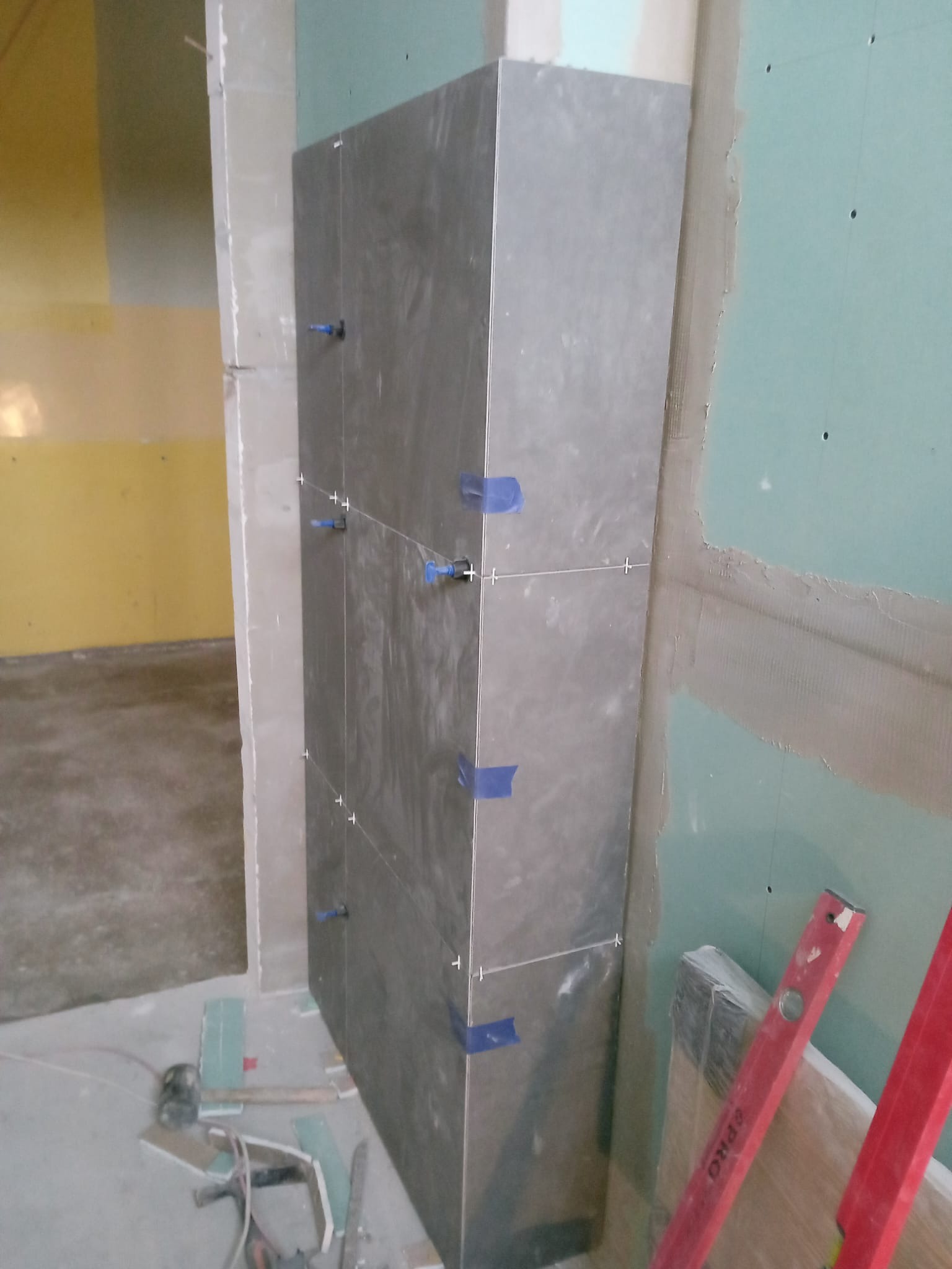 Fragment ściany obłożonej ciemnymi płytkami, zabezpieczonymi niebieską taśmą i krzyżykami dystansowymi, obok ściana z płyty gipsowo-kartonowej, na podłodze narzędzia i resztki płytek.