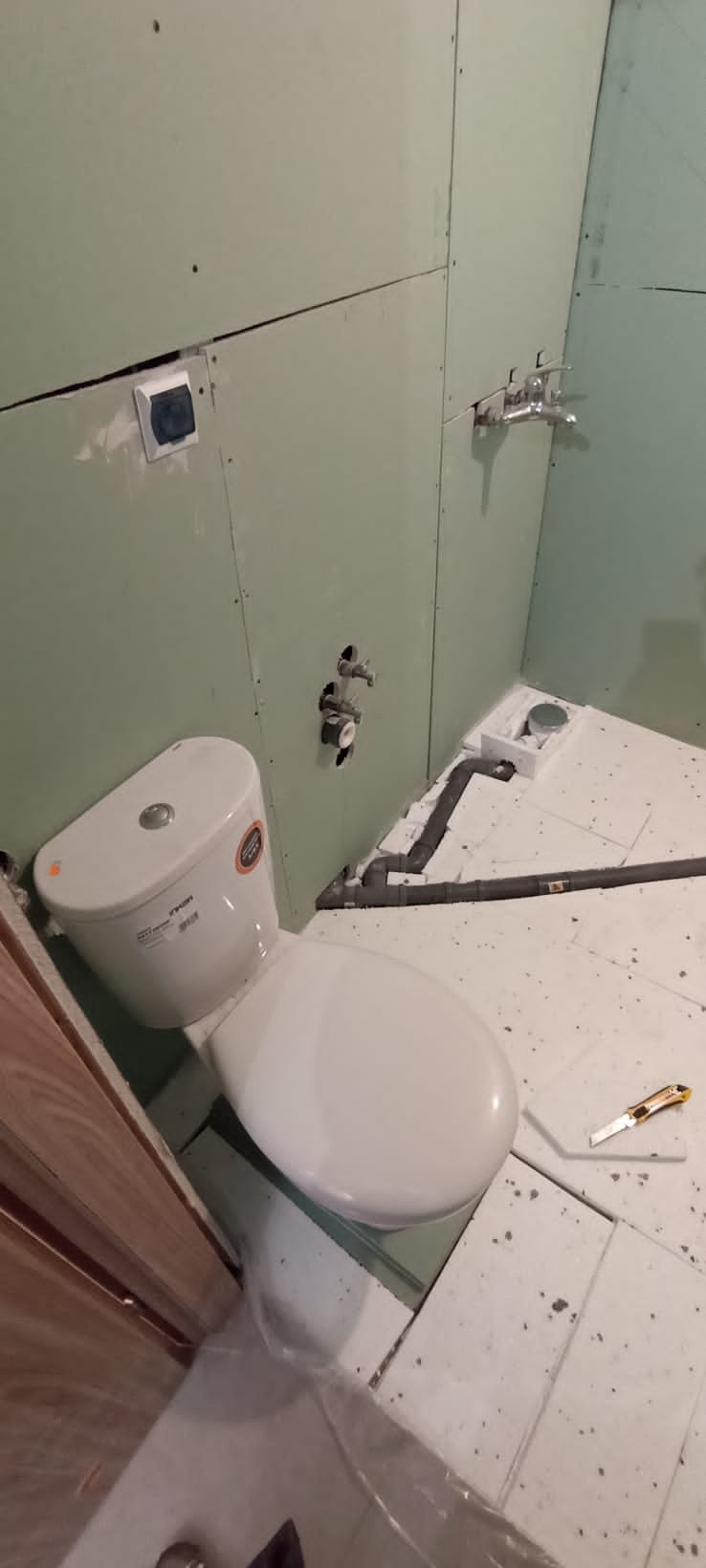 Niedokończona łazienka: biała toaleta z naklejką producenta, częściowo położone płytki podłogowe z widocznym nożem do cięcia, ściany wyłożone płytami gipsowo-kartonowymi z widocznymi instalacjami...