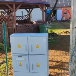 Nowo zainstalowana, szara skrzynka elektryczna z oznaczeniami ostrzegawczymi w Kielnie, na tle altany, domu z panelami słonecznymi i minikoparki.